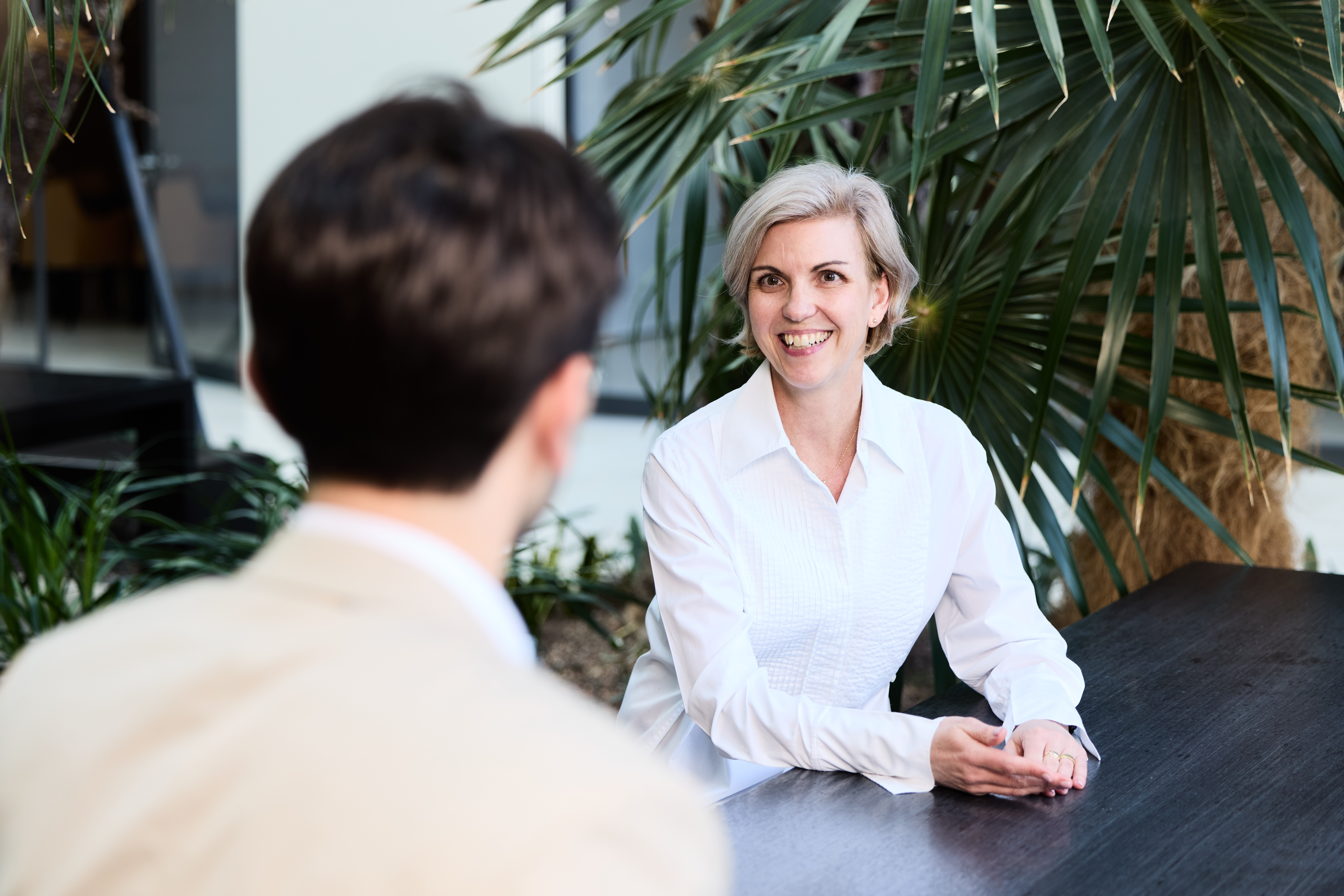 Eine Frau mit kurzem blondem Haar sitzt in einem hellen Innenbereich mit Pflanzen, lächelt freundlich und spricht mit einer gegenüber sitzenden Person bei der Baader Bank.