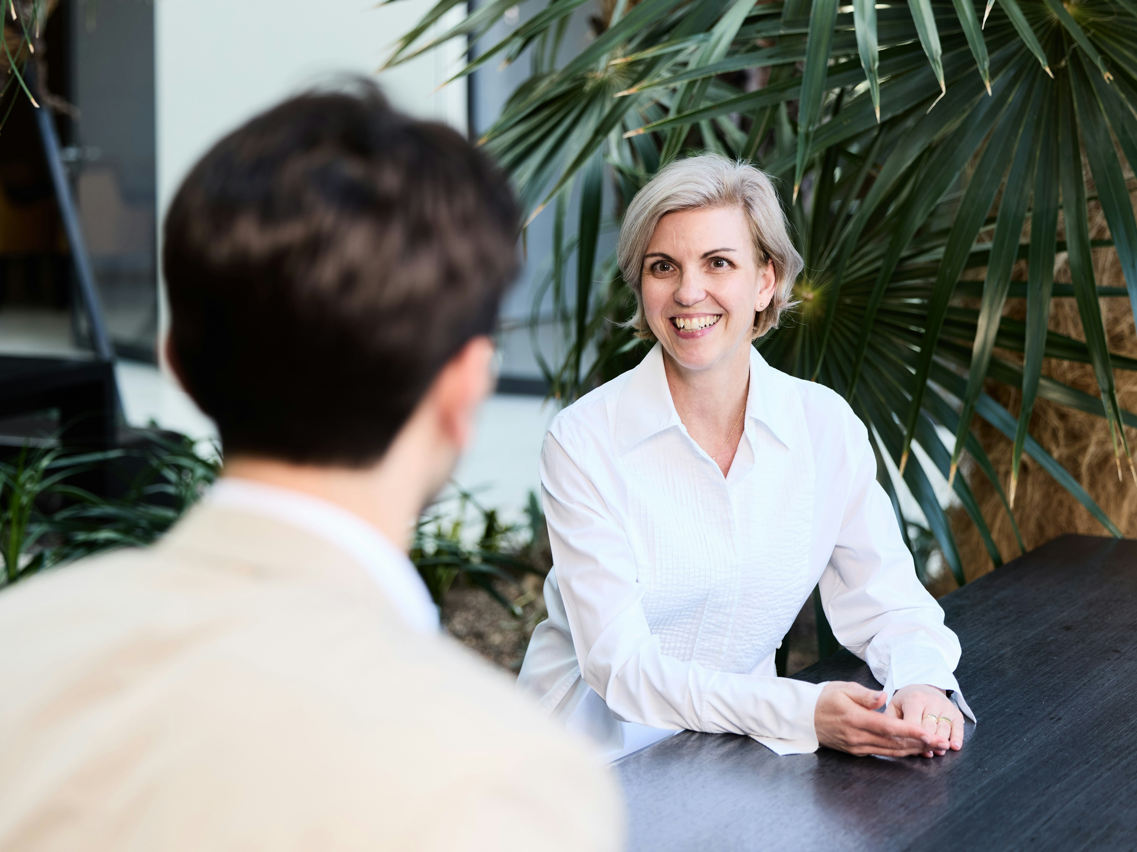Eine Frau mit kurzem blondem Haar sitzt in einem hellen Innenbereich mit Pflanzen, lächelt freundlich und spricht mit einer gegenüber sitzenden Person bei der Baader Bank.