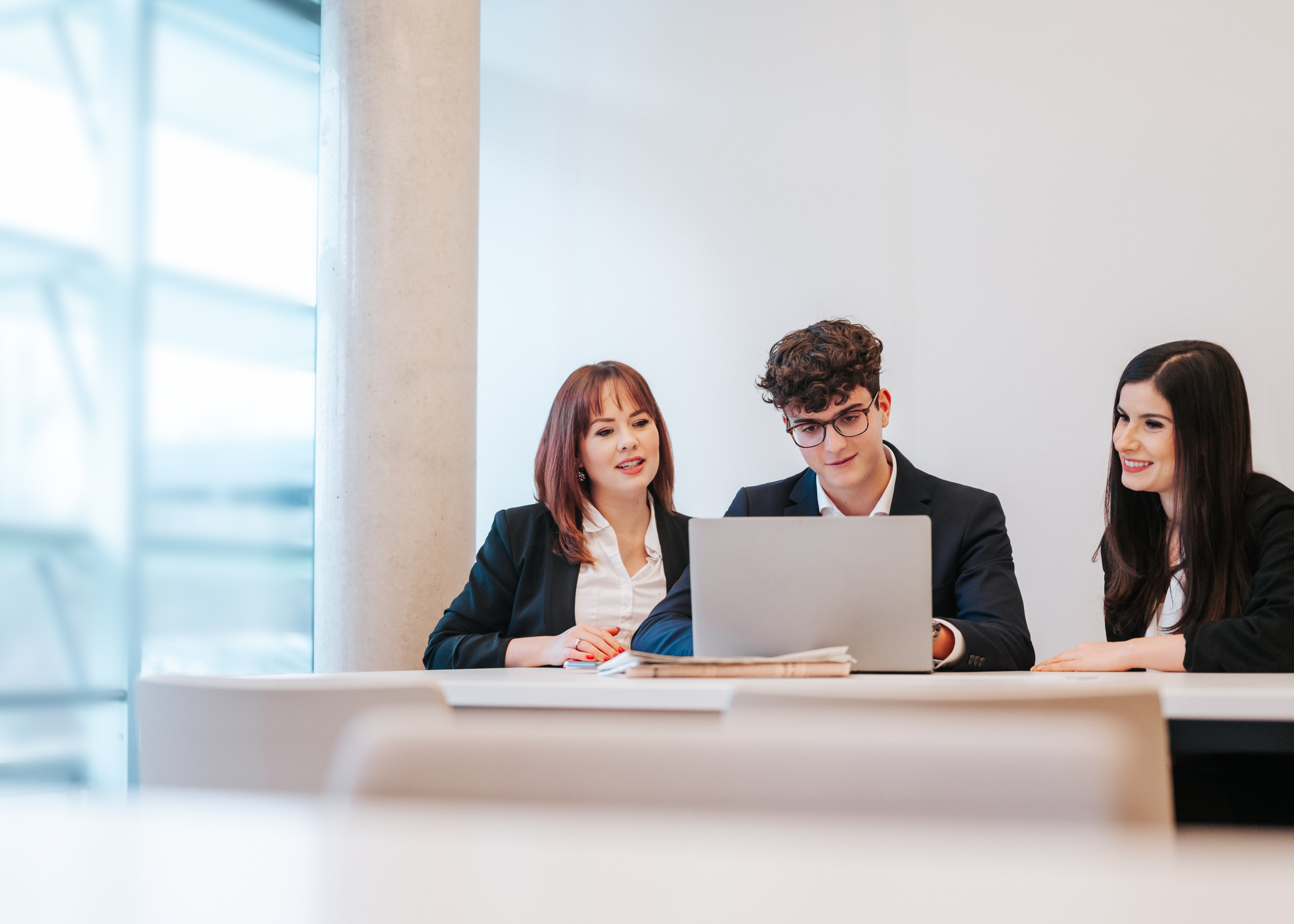 Zwei Frauen und ein Mann im Business-Outfit sitzen nebeneinander an einem Tisch und arbeiten am Laptop.