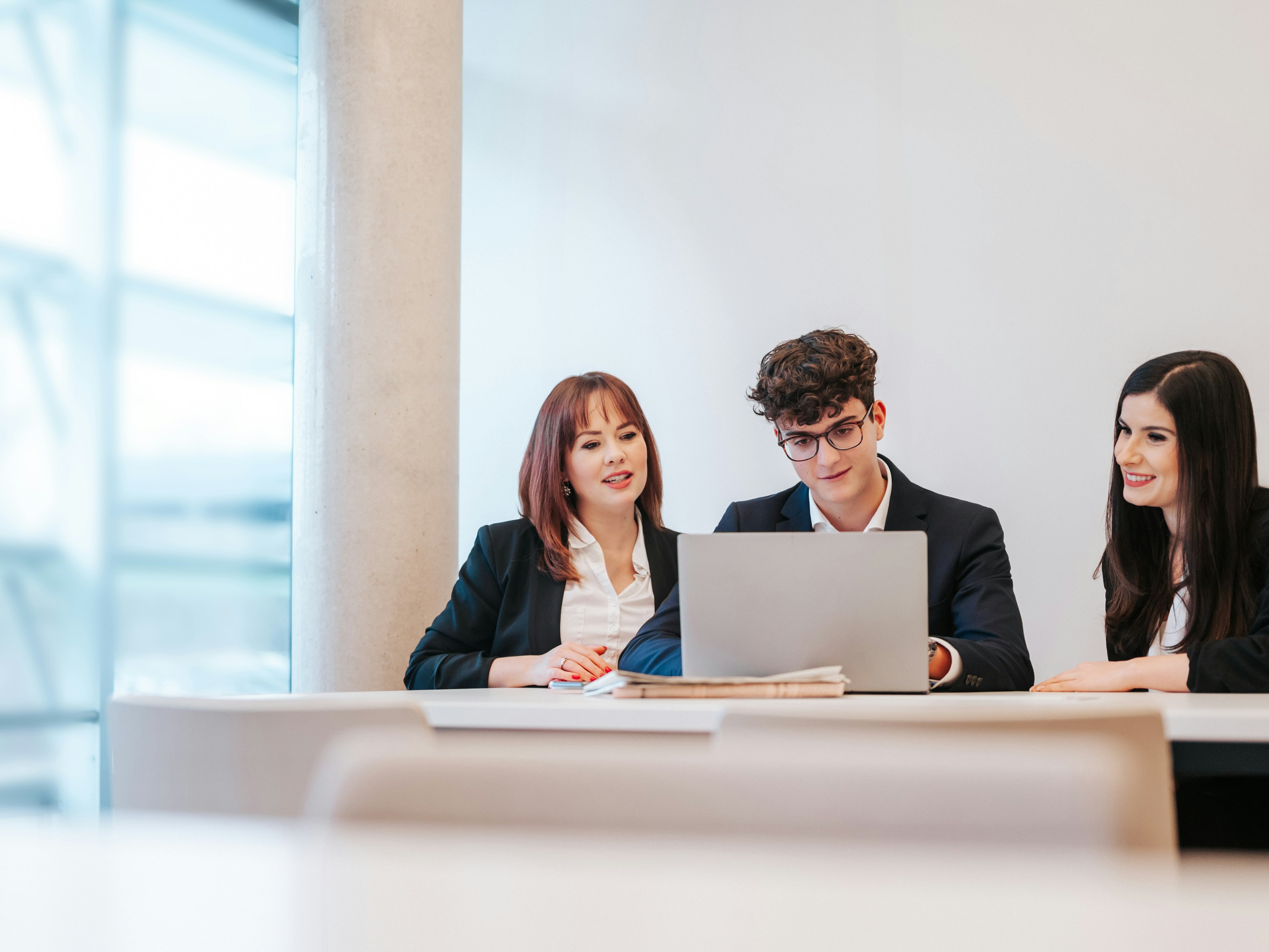 Zwei Frauen und ein Mann im Business-Outfit sitzen nebeneinander an einem Tisch und arbeiten am Laptop.