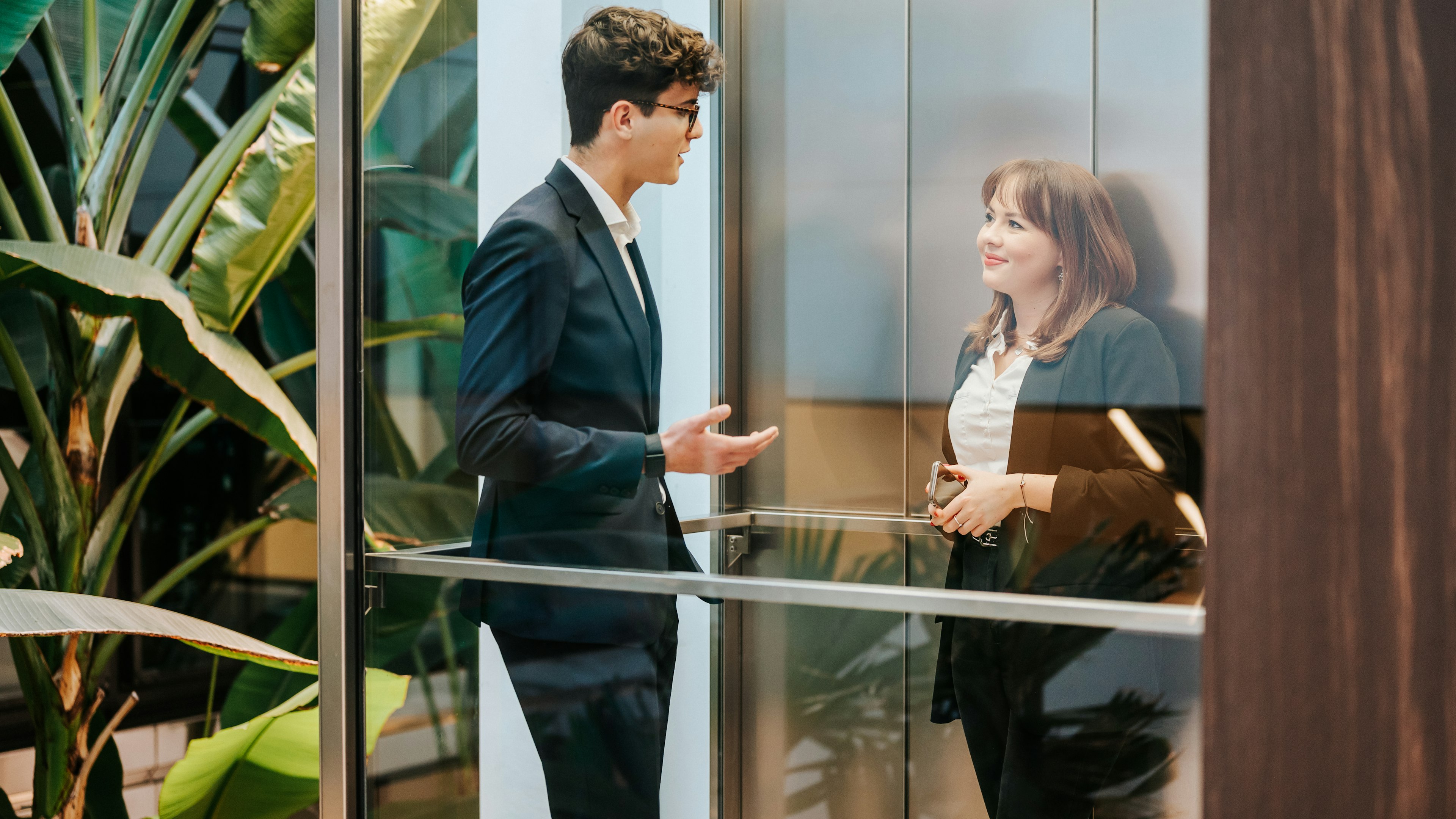 Zwei Baaderbank Mitarbeiter im Anzug im Aufzug, Gesprächsszene im modernen Atrium mit Pflanzen.