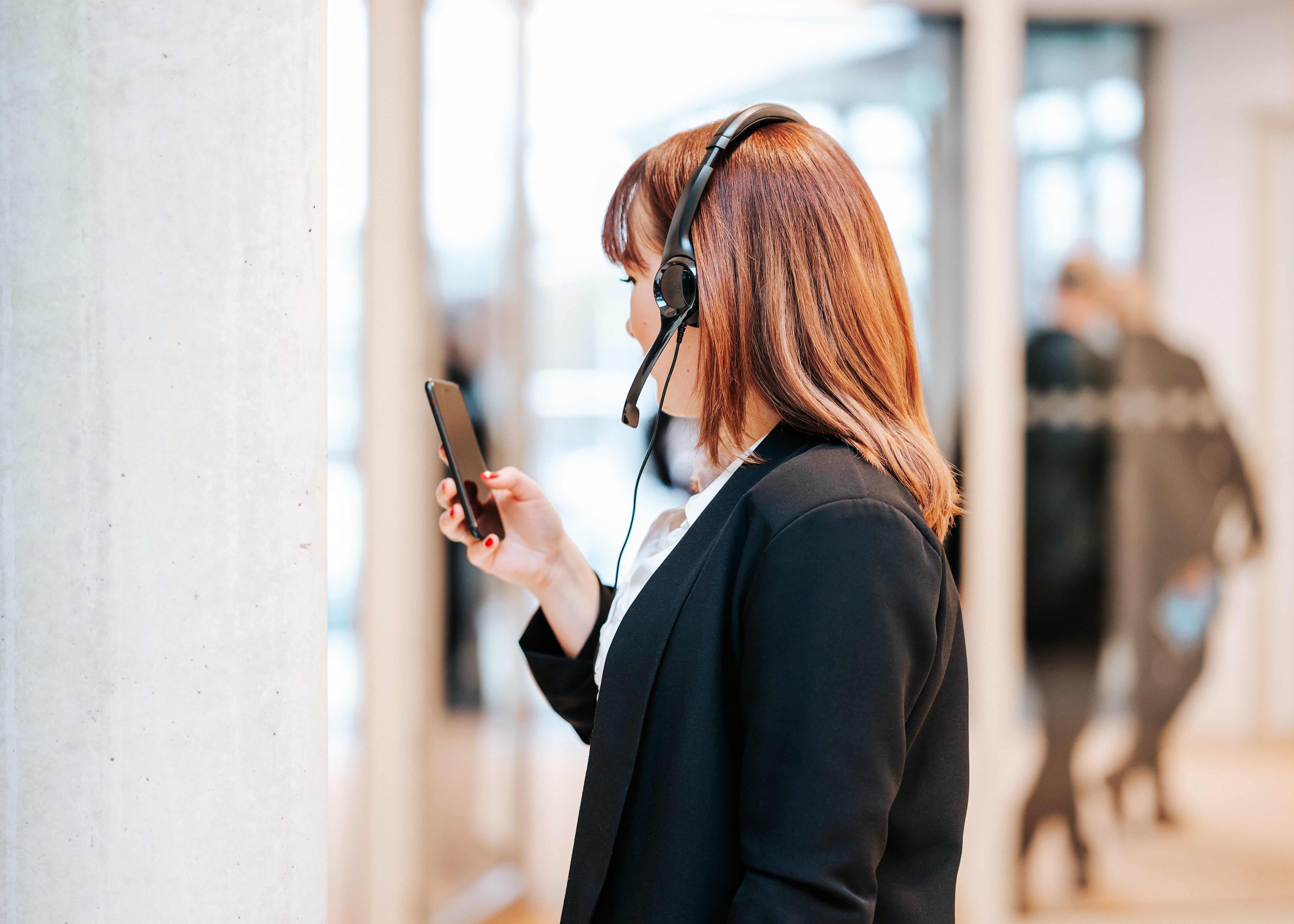 Frau mit braunen Haaren und Headset, trägt schwarzen Blazer, schaut im Büro auf ihr Smartphone.