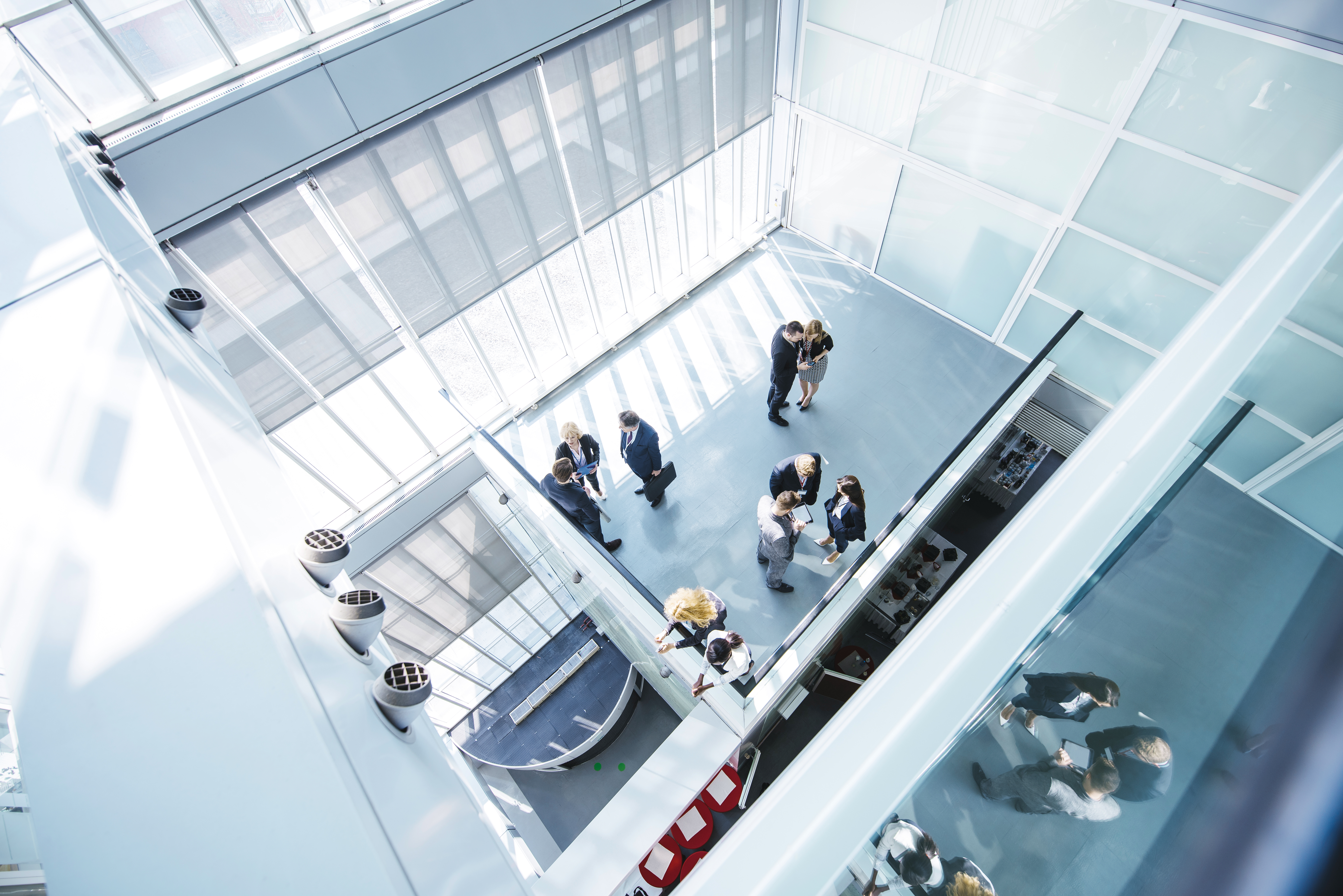 Blick von oben auf eine Gruppe von Menschen in Businesskleidung, die sich in einem modernen, lichtdurchfluteten Atrium unterhalten. Das Glas der Architektur spiegelt einige der Personen.