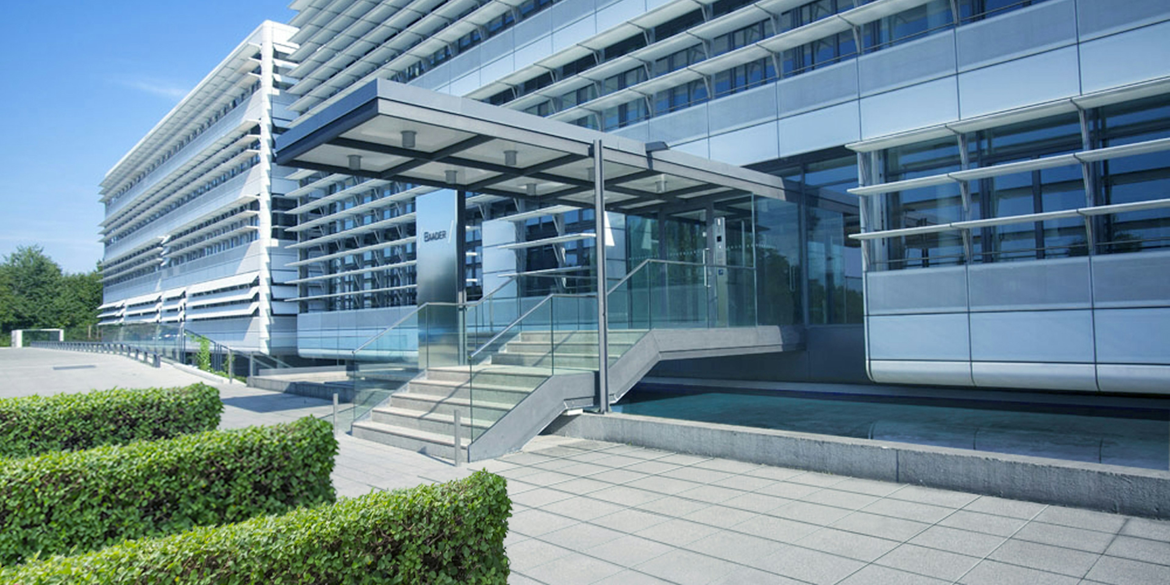 Moderner Eingangsbereich der Baader Bank Zentrale mit Glasfassade, breiter Treppe und klaren architektonischen Linien bei sonnigem Wetter.