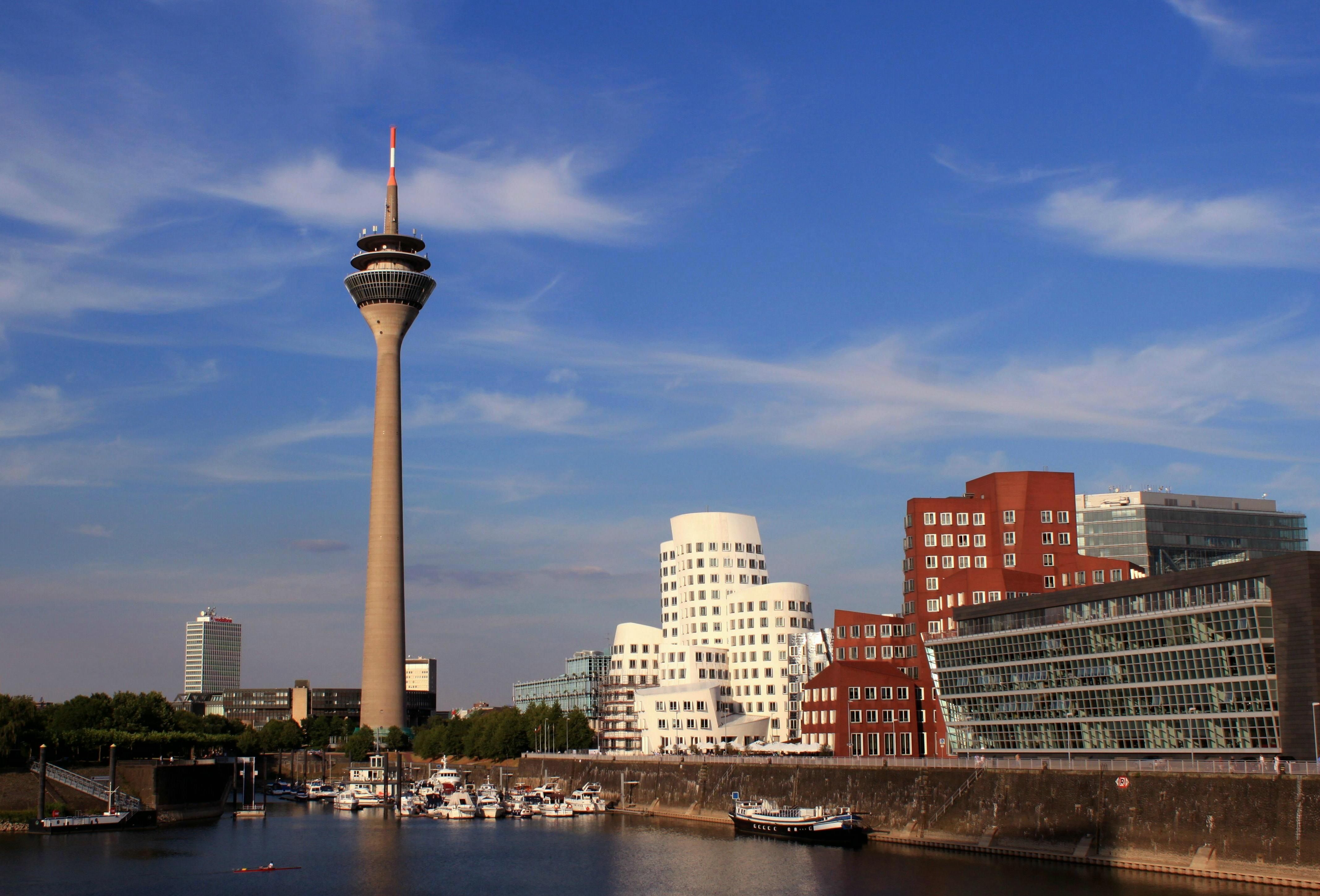 Blick auf den Düsseldorfer Medienhafen mit dem Rheinturm links, den weißen und roten Gehry-Bauten in der Mitte sowie weiteren modernen Gebäuden am Wasser unter blauem Himmel.