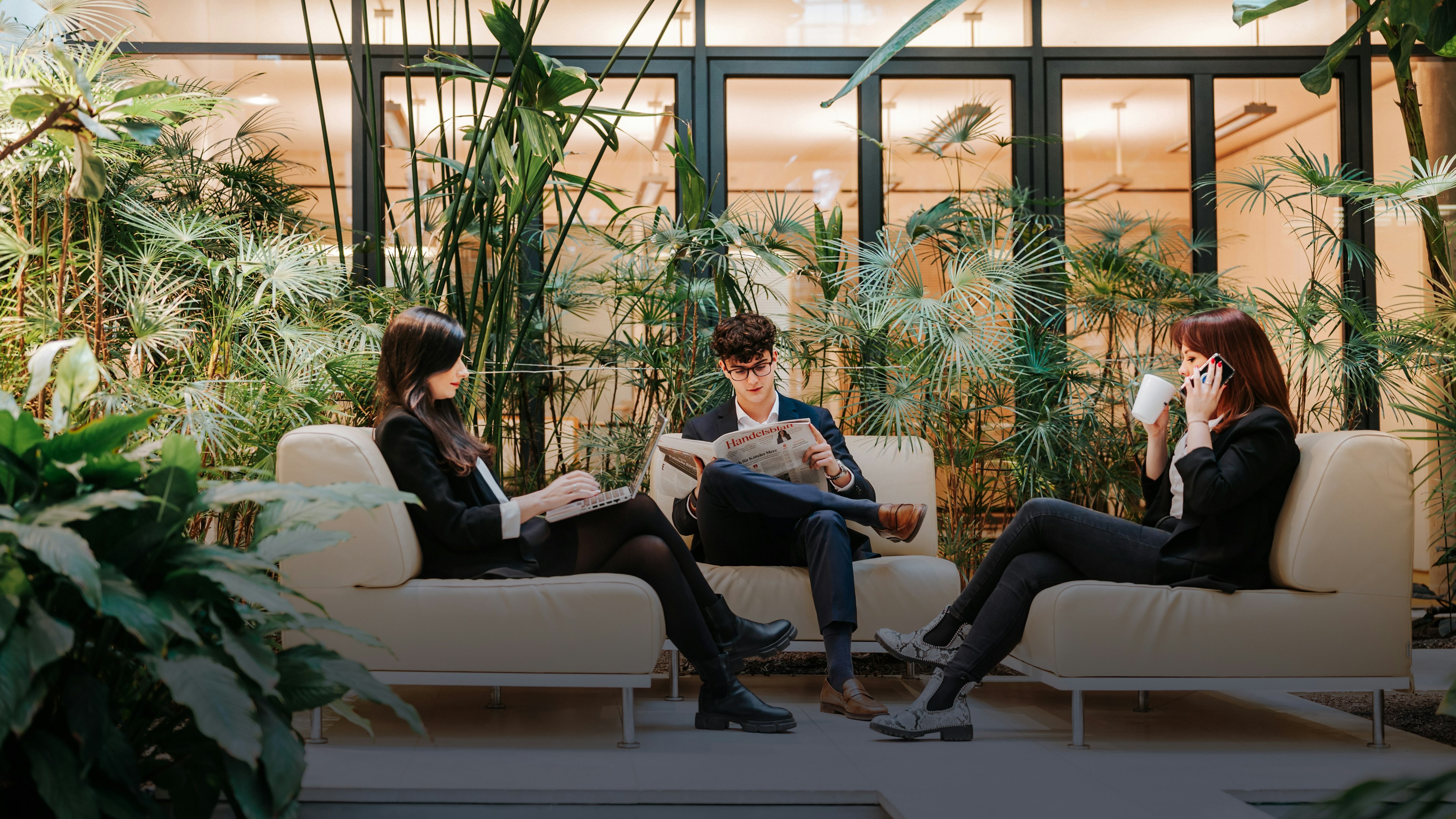 Drei Mitarbeitende der Baaderbank sitzen in einer begrünten Lounge mit Zeitung, Laptop und Kaffeetasse.