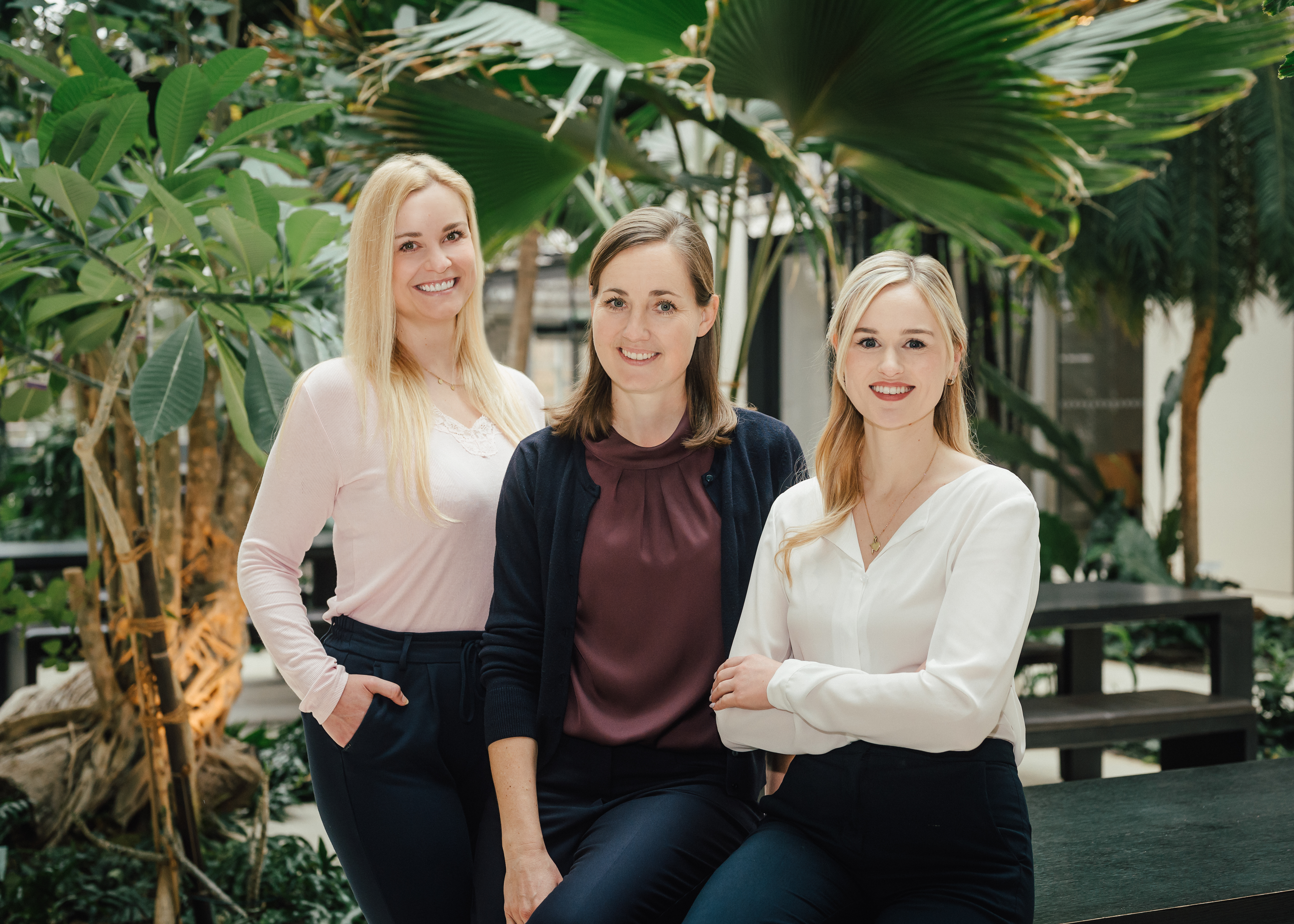 rei Frauen stehen nebeneinander in einem begrünten Innenbereich, sie tragen ein rosa Oberteil, ein weinrotes Oberteil mit dunkelblauer Strickjacke und eine weiße Bluse.