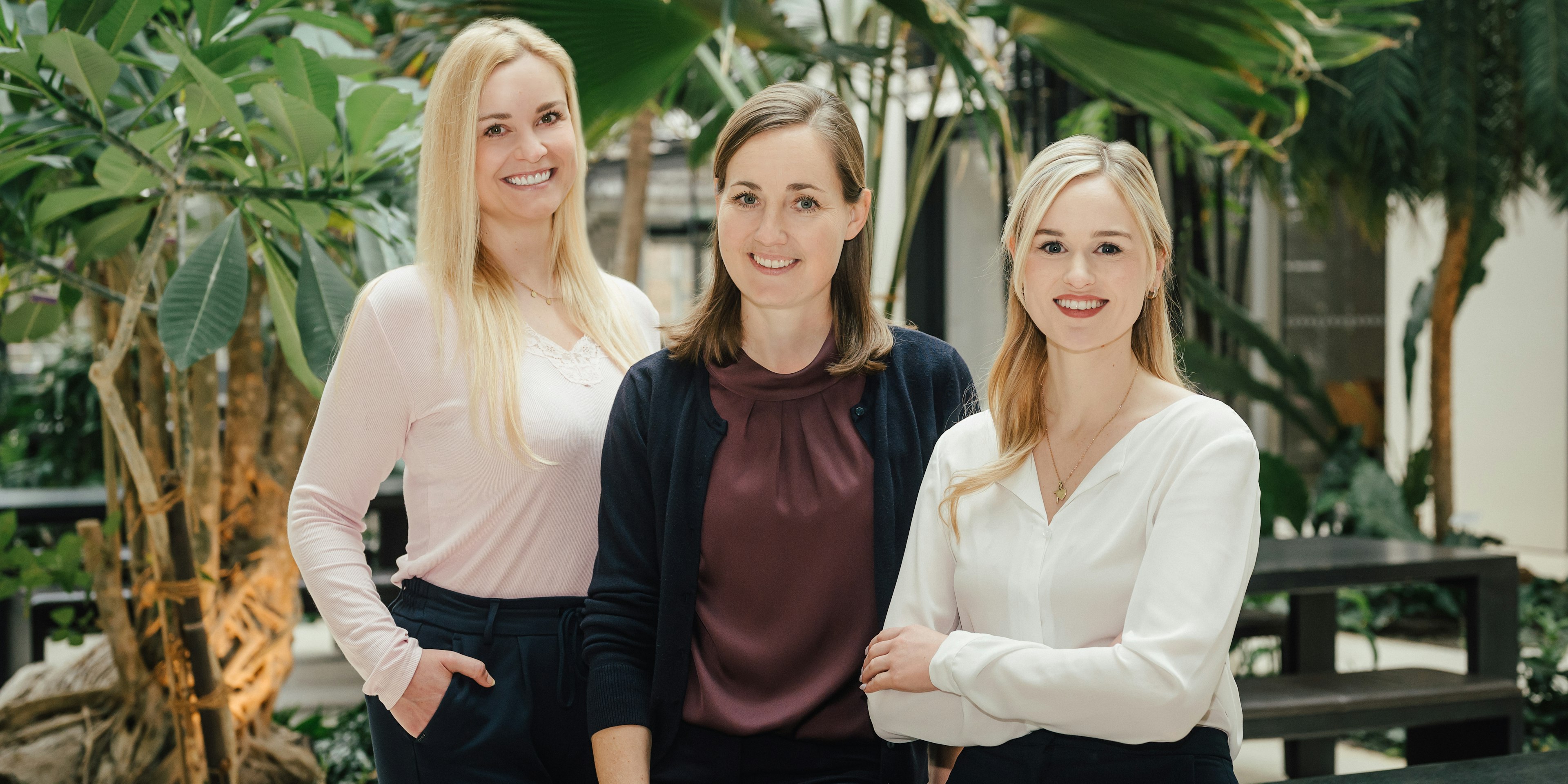 rei Frauen stehen nebeneinander in einem begrünten Innenbereich, sie tragen ein rosa Oberteil, ein weinrotes Oberteil mit dunkelblauer Strickjacke und eine weiße Bluse.