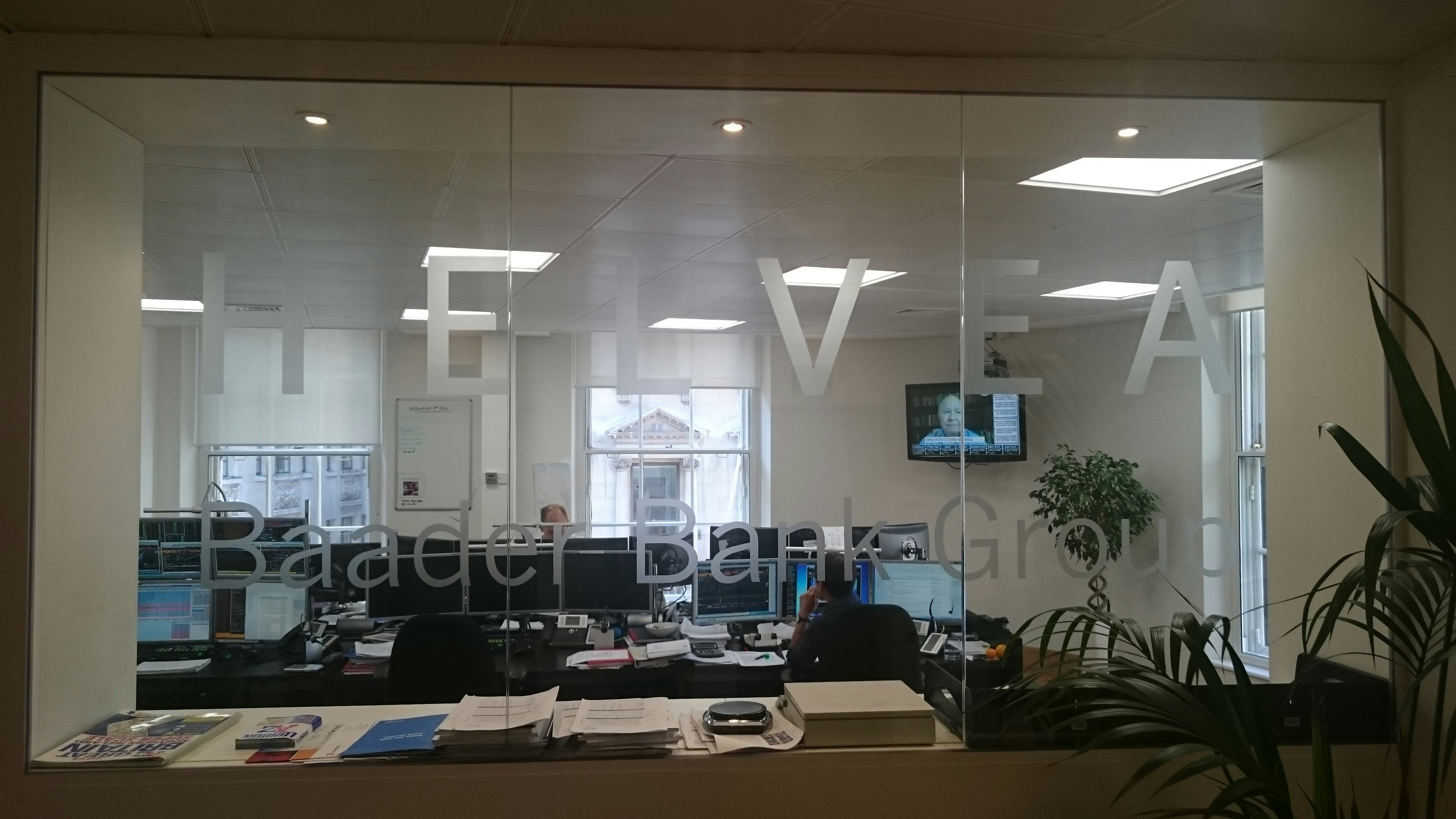 Glaswand mit Aufschrift "Baader Bank Group" im Londoner Büro der Baaderbank, dahinter Arbeitsplätze.