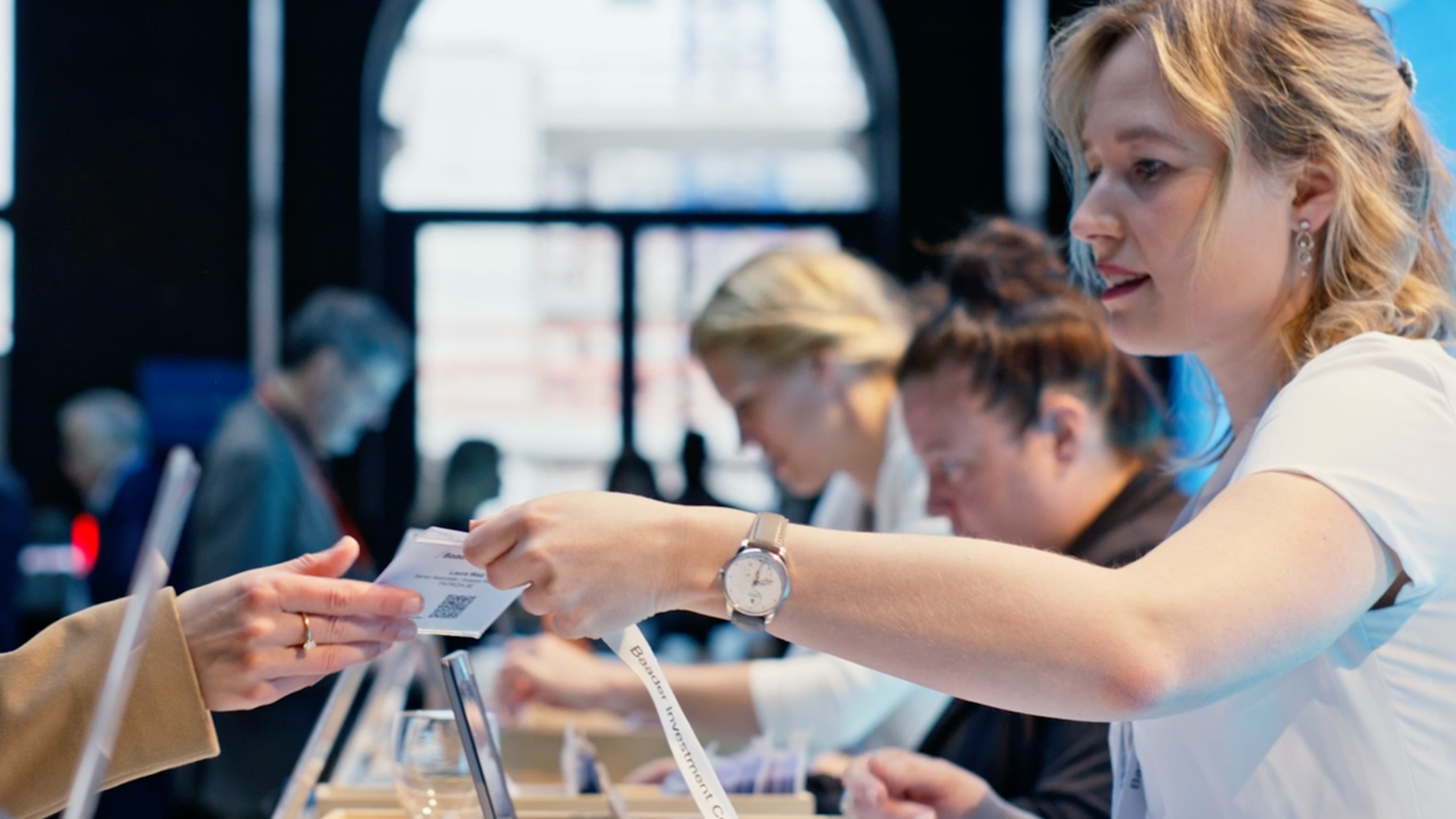 Mitarbeiterin der Baaderbank übergibt ein Ticket an eine Person am Empfangstisch.