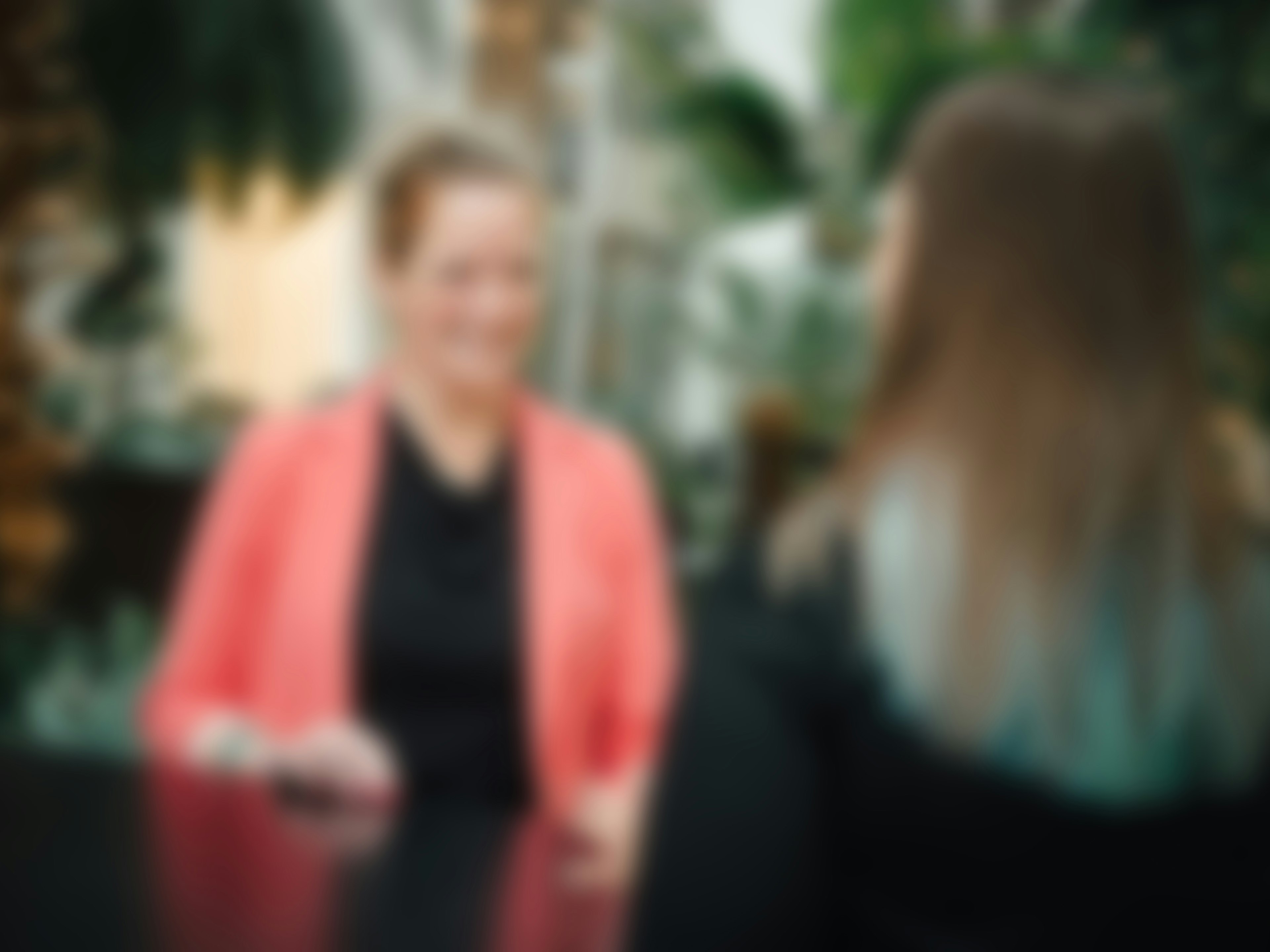 Unscharfe Aufnahme von zwei Frauen an einem Tisch, im Hintergrund sind große grüne Pflanzen erkennbar.