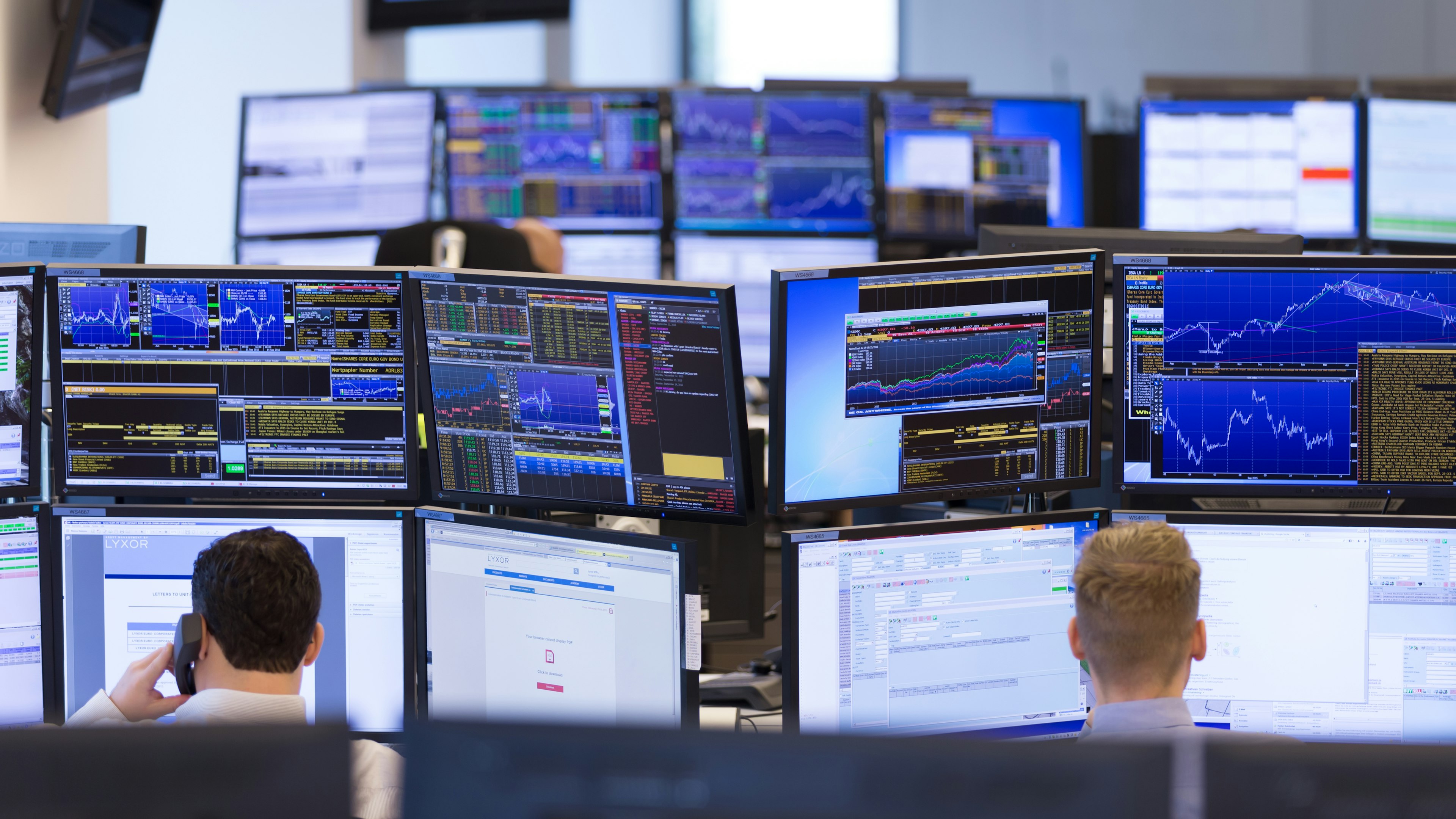 Arbeitsplätze mit vielen Monitoren, auf den Bildschirmen sind Finanzdaten und Kursgrafiken zu sehen.