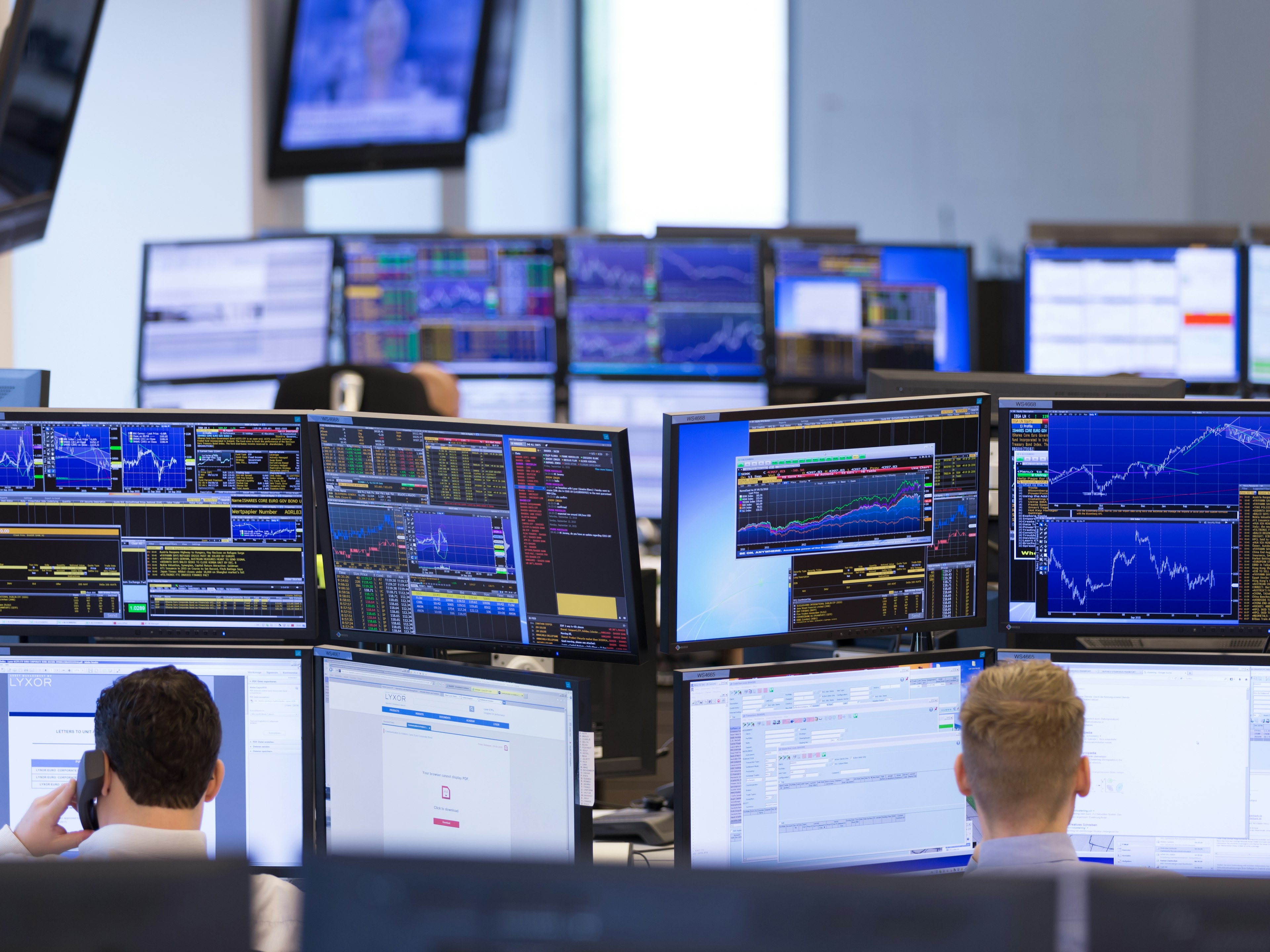 Arbeitsplätze mit vielen Monitoren, auf den Bildschirmen sind Finanzdaten und Kursgrafiken zu sehen.