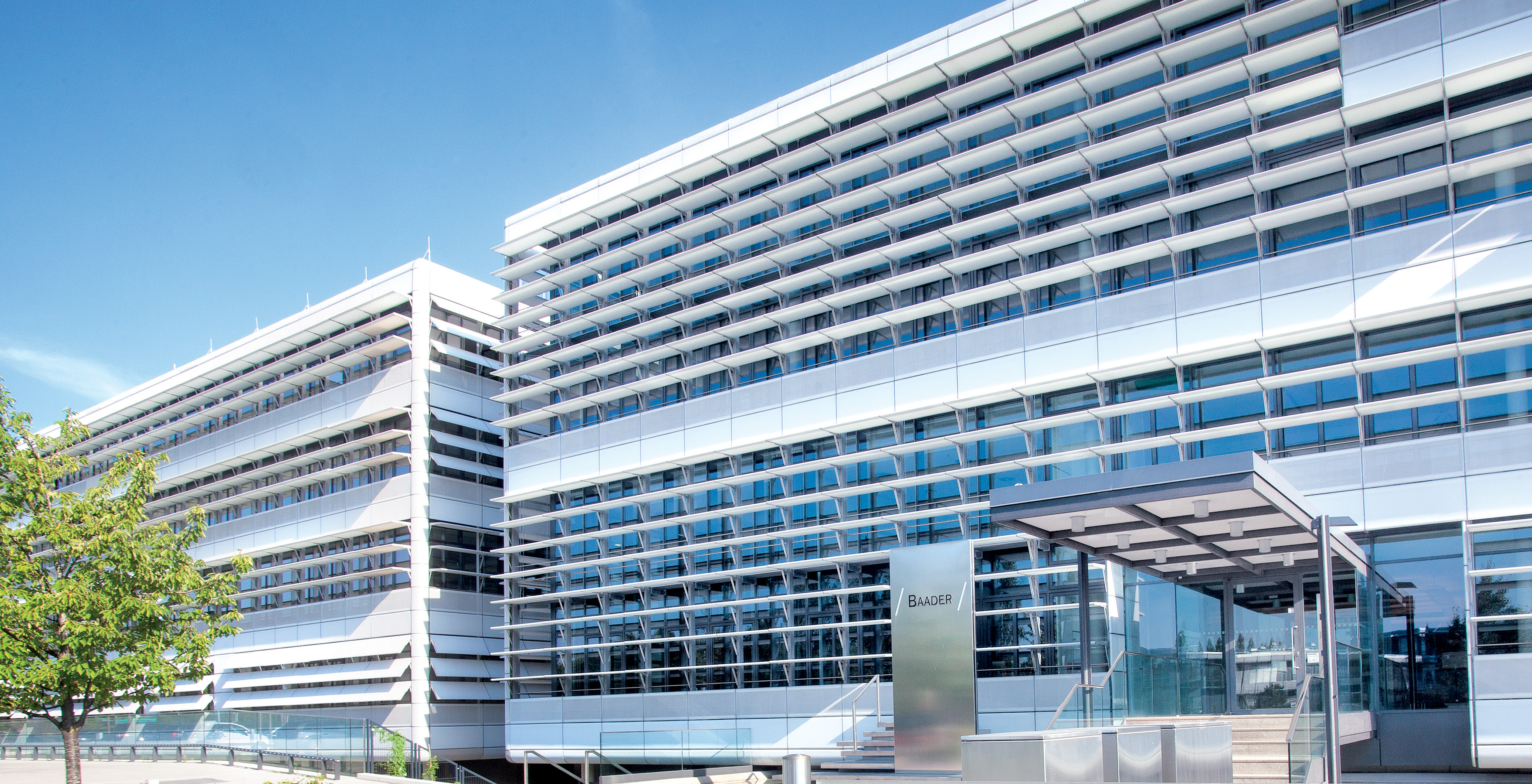 Moderne Baaderbank Zentrale in Unterschleißheim mit Glasfassade, Eingang und Baum im Vordergrund.