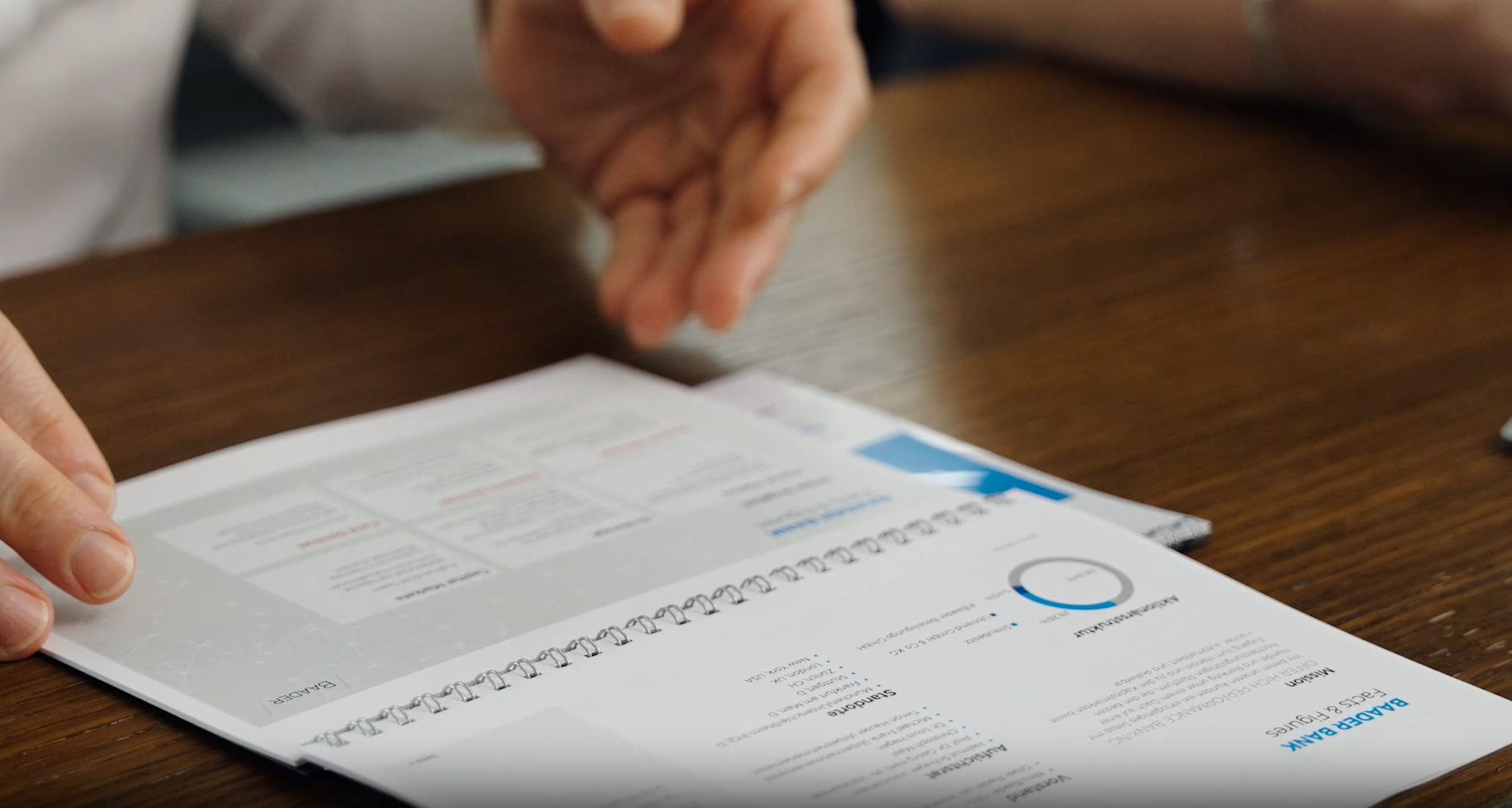 uf einem Holztisch liegt ein aufgeschlagenes Ringbuch mit dem Titel „Baader Bank Facts & Figures“, daneben eine ausgestreckte Hand.