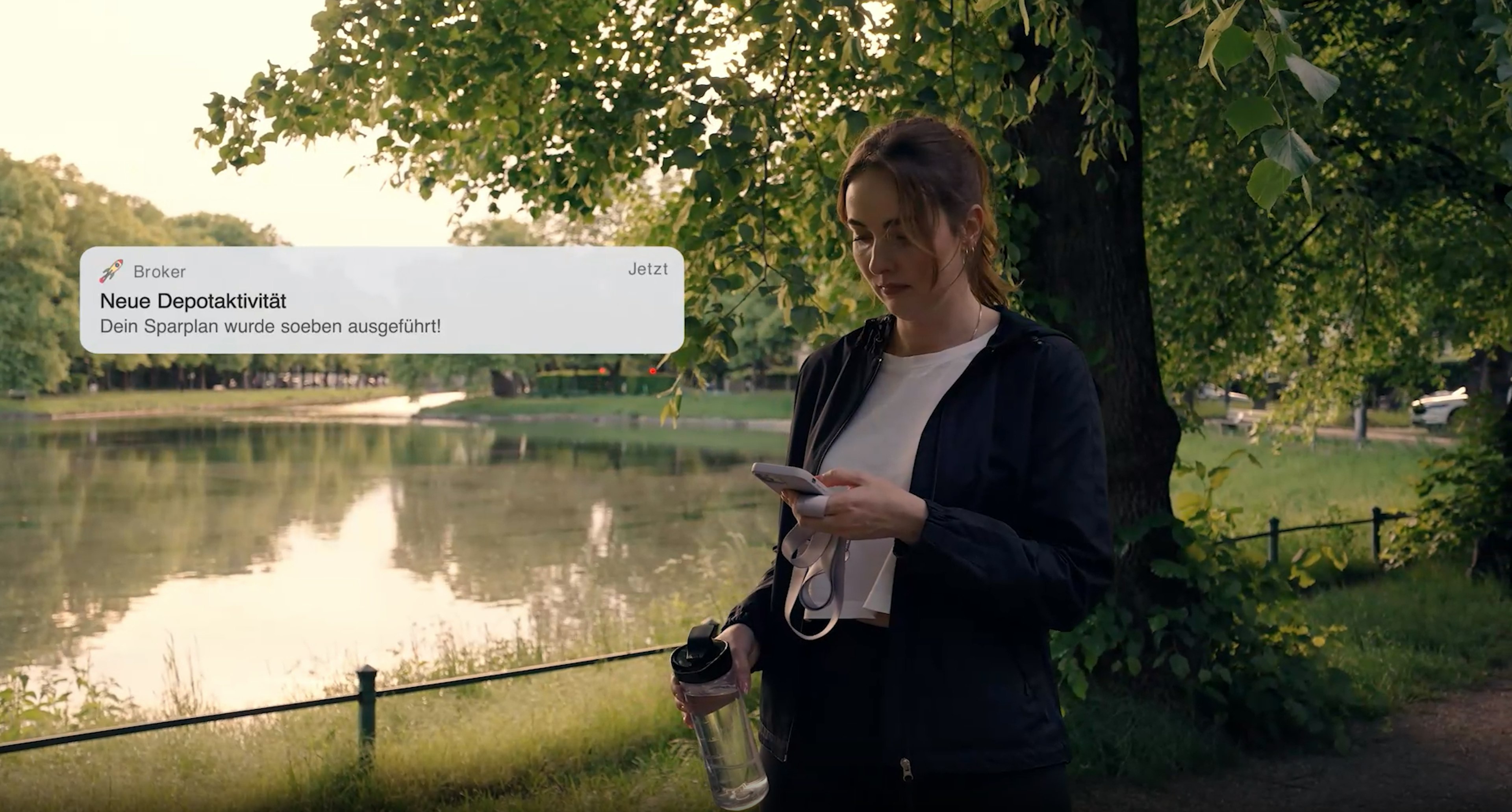 Eine Frau steht in Sportkleidung mit Trinkflasche und Smartphone in der Hand in einem Park. Auf ihrem Handy erscheint eine Push-Benachrichtigung mit dem Text „Neue Depotaktivität – Dein Sparplan wurde soeben ausgeführt!“.