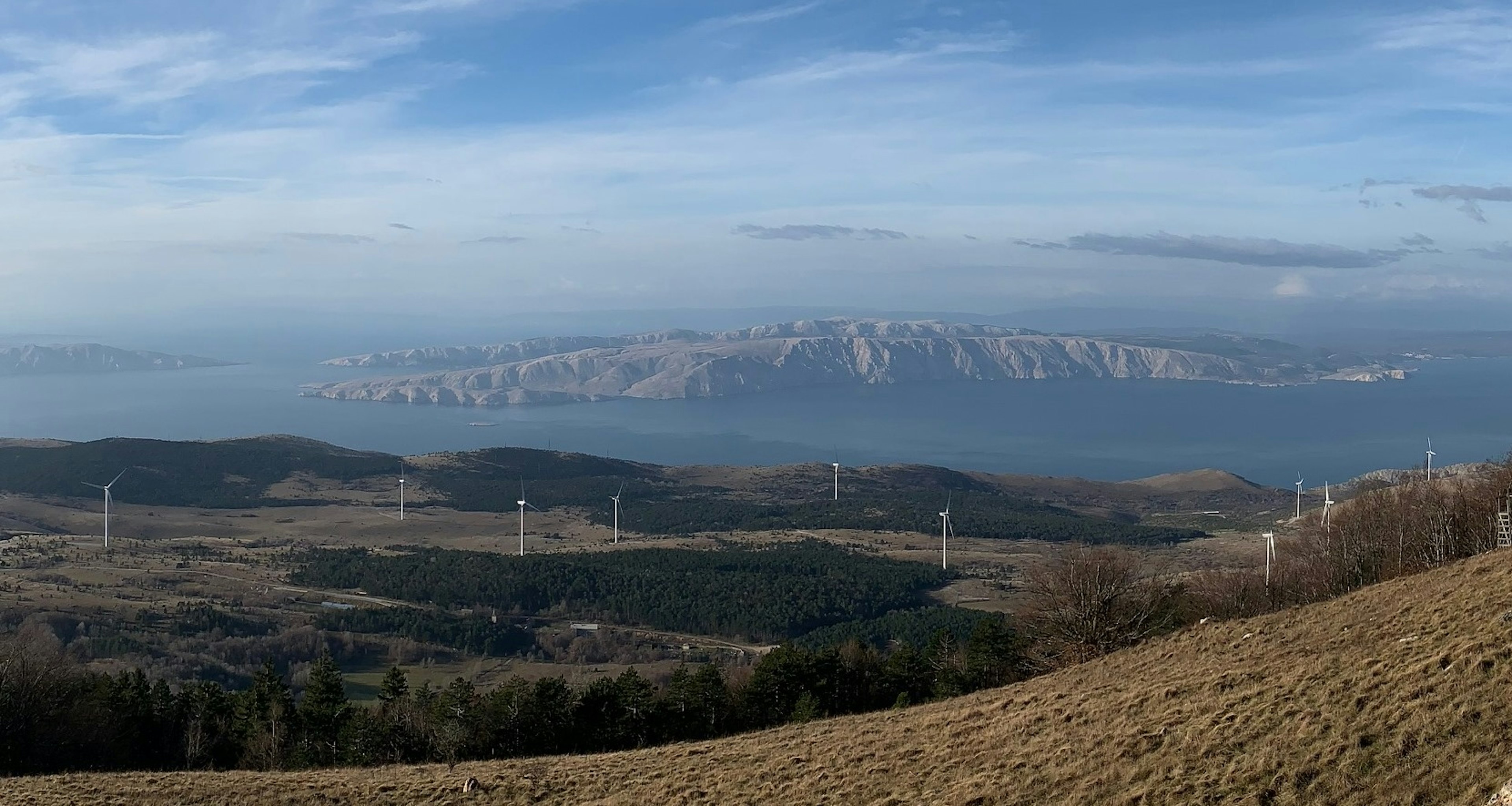 Landschaft mit mehreren Windrädern, dahinter Meer und felsige Inseln unter blauem Himmel.