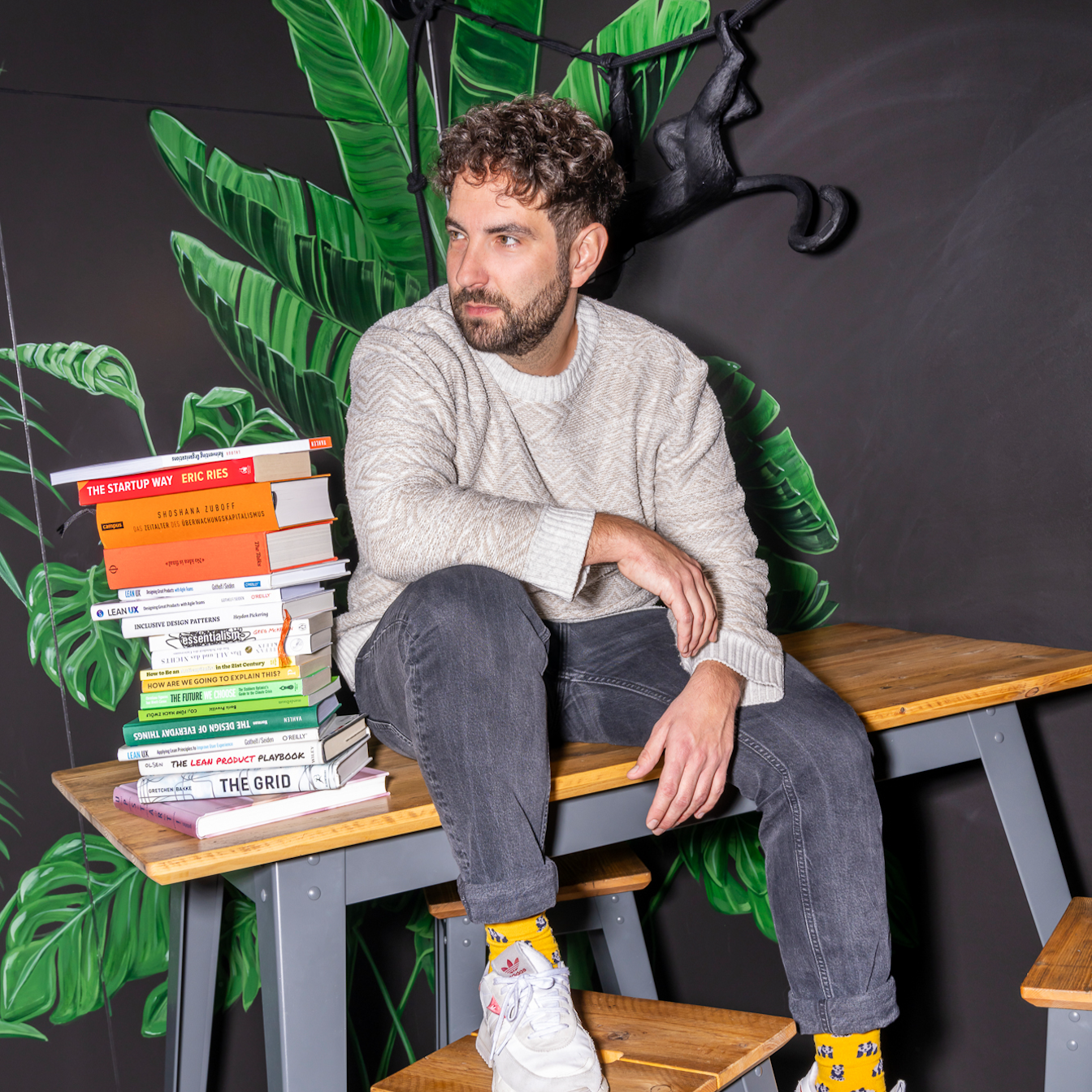 Ein Mann in einem grauen Pullover und Jeans sitzt auf einem Holztisch neben einem Stapel Bücher, im Hintergrund ist ein Wandgemälde mit Dschungelmotiven zu sehen.
