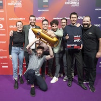 Eine Gruppe von acht Personen posiert gemeinsam auf einem violetten Teppich und hält eine goldene Trophäe in Raketenform und eine Plakette mit der Aufschrift „Best of Web Technology Gold 2024“.