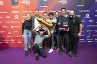 Eine Gruppe von acht Personen posiert gemeinsam auf einem violetten Teppich und hält eine goldene Trophäe in Raketenform und eine Plakette mit der Aufschrift „Best of Web Technology Gold 2024“.