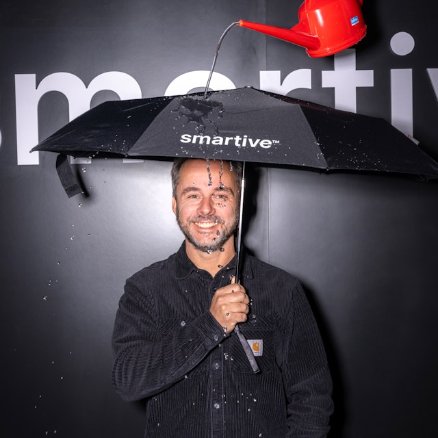 Ein Mann steht unter einem "intelligenten" Regenschirm, während Wasser aus einer roten Giesskanne austritt und ihn durchnässt.