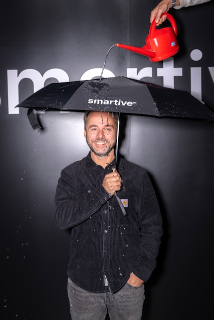 Ein Mann steht unter einem "intelligenten" Regenschirm, während Wasser aus einer roten Giesskanne austritt und ihn durchnässt.