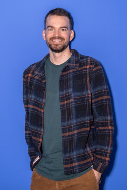 Mann mit kurzen Haaren und Bart in grünem Hemd, karierter Flanelljacke und brauner Hose steht lächelnd vor einem einfarbig blaün Hintergrund.