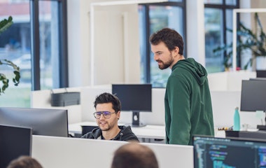 Zwei Entwickler in einem modernen Büro, einer sitzend vor dem Monitor, der andere stehend daneben und zuschauend.