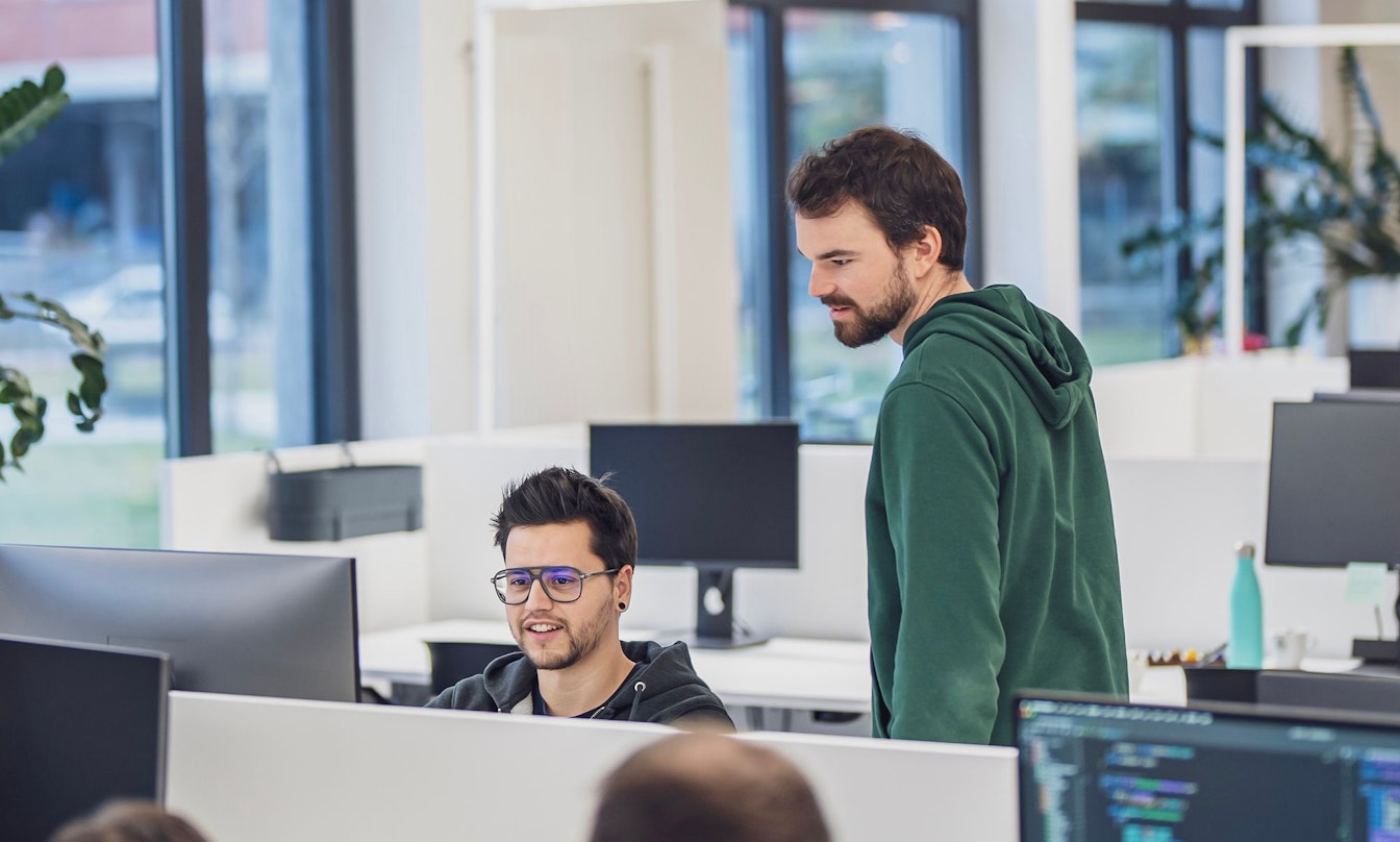 Zwei Entwickler in einem modernen Büro, einer sitzend vor dem Monitor, der andere stehend daneben und zuschauend.
