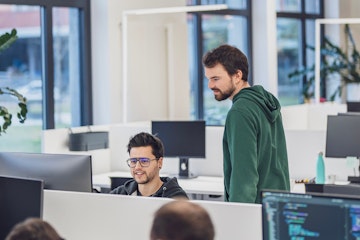 Zwei Entwickler in einem modernen Büro, einer sitzend vor dem Monitor, der andere stehend daneben und zuschauend.