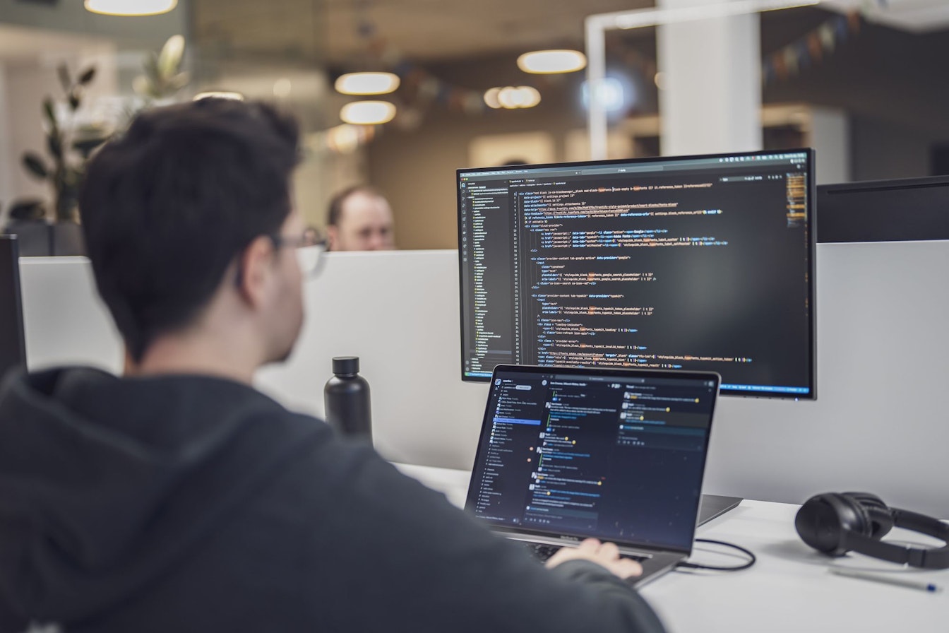 Person mit Brille, die an einem Laptop arbeitet. Auf dem Laptop-Bildschirm und einem grösseren Monitor werden Codezeilen angezeigt. Auf dem Schreibtisch sind eine Wasserflasche, Kopfhörer und eine Pflanze zu sehen.