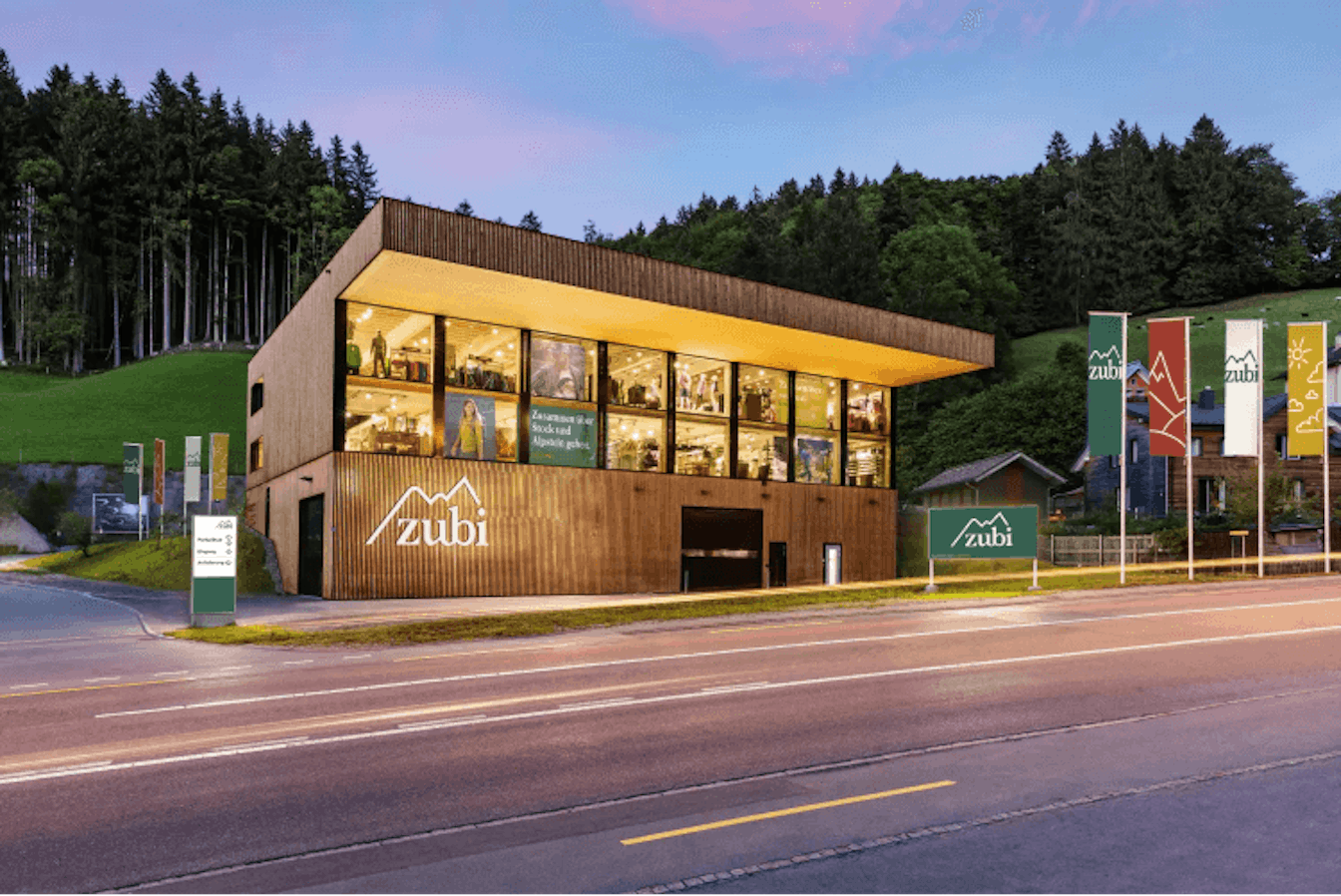 Ein modernes Holzgebäude mit grossen Fenstern und „Zubi“-Schildern steht neben einer Straße, umgeben von üppigem Grün und bewaldeten Hügeln. Davor sind Flaggen mit verschiedenen Motiven zu sehen.
