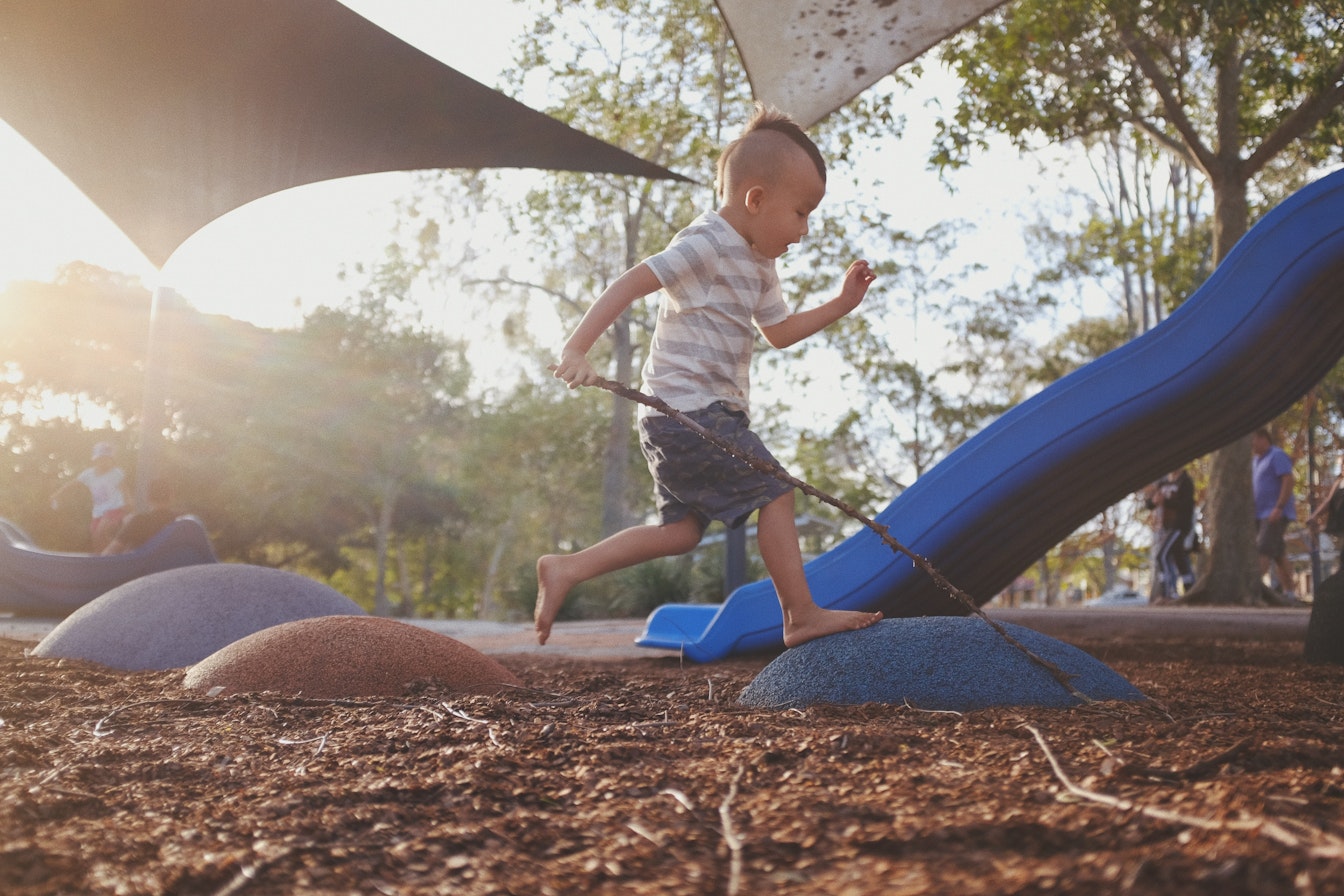 Ein Kind im gestreiften Hemd springt mit einem Stock in der Hand über einen blauen Hügel auf einem Spielplatz. Im Hintergrund sind eine Rutsche und Schatten spendende Strukturen zu sehen.