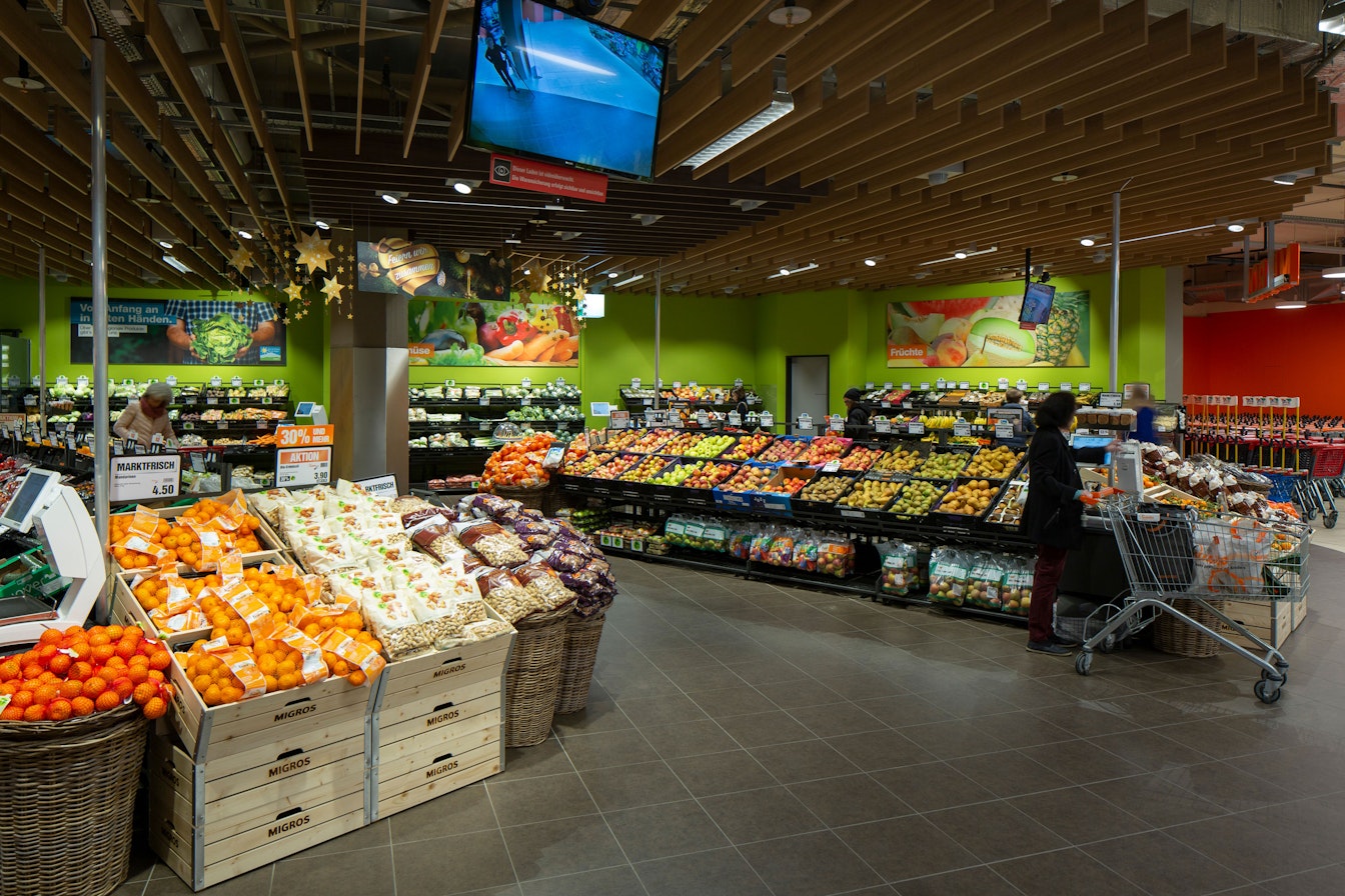 Die Obst- und Gemüseabteilung eines Lebensmittelladens mit verschiedenen Obst- und Gemüsesorten, die auf Holzkisten und Regalen präsentiert werden. Käufer mit Einkaufswagen sind da und ein Fernsehbildschirm hängt von der Decke.