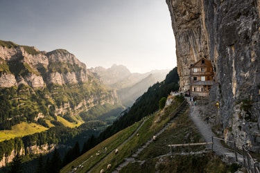 Ein Holzhaus mit felsiger Aussenfassade ist in einen steilen Berghang gebaut. Umgeben von üppigem Grün und einer fernen Berglandschaft grasen Schafe auf dem abschüssigen Gelände.