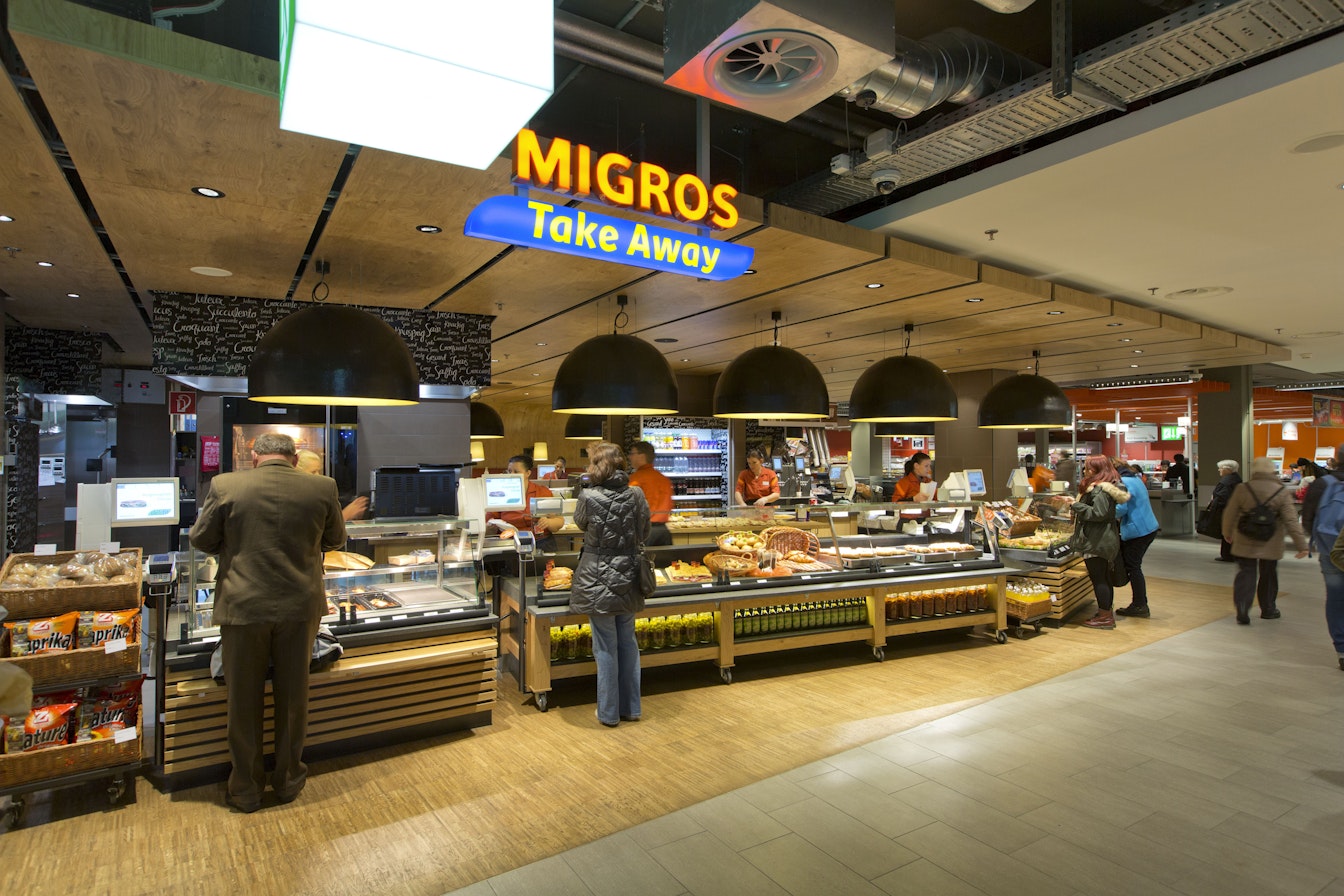Kunden bestellen Essen an der Migros-Take-Away-Theke in einem Einkaufszentrum. Verschiedene Lebensmittel werden unter hellem Licht präsentiert.