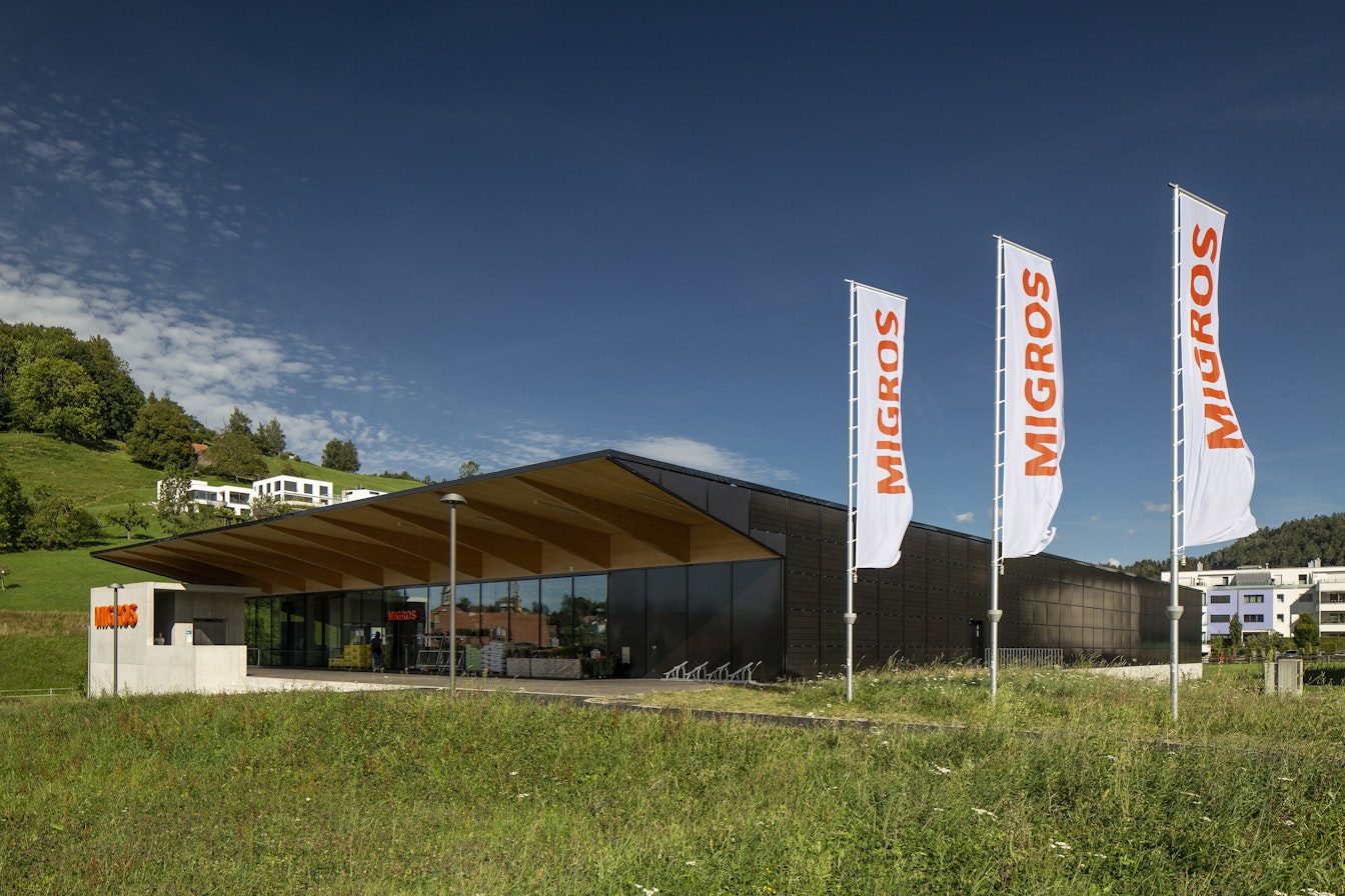 Ein modernes Gebäude mit Holzdach und grossen Glasfenstern, mit mehreren „MIGROS“-Flaggen vor einer hügeligen Landschaft und einem blauen Himmel.