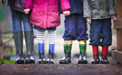 Eine Gruppe Kinder steht in einer Reihe. Sie tragen bunte Gummistiefel mit Schlamm darauf. Nur ihre Beine und Stiefel sind zu sehen.