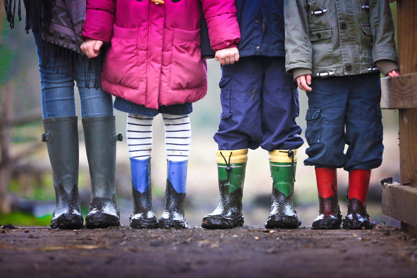 Eine Gruppe Kinder steht in einer Reihe. Sie tragen bunte Gummistiefel mit Schlamm darauf. Nur ihre Beine und Stiefel sind zu sehen.