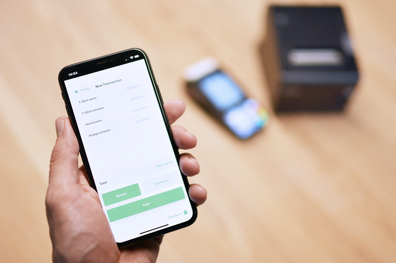 Nahaufnahme einer Person, die ein Smartphone hält, auf dem der Bildschirm einer Zahlungs-App neben einem unscharfen Kartenleser und einem kleinen schwarzen Kästchen auf einer Holzoberfläche angezeigt wird.