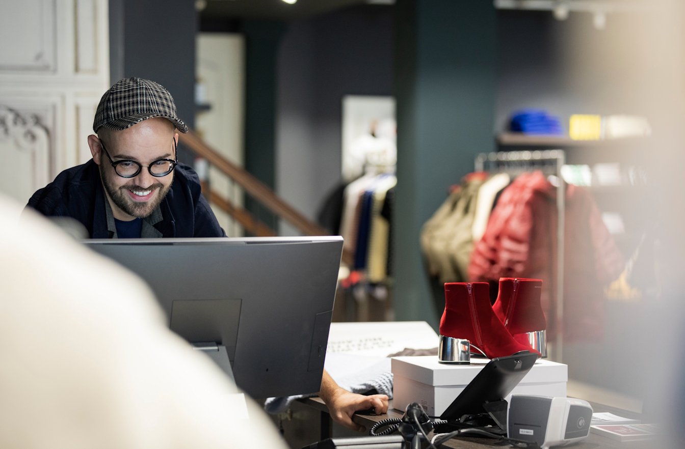 Ein Verkäufer mit Brille und Kappe lächelt und arbeitet an einem Computer in einem Bekleidungsgeschäft, während auf dem Tresen rote Stiefeletten ausgestellt sind.