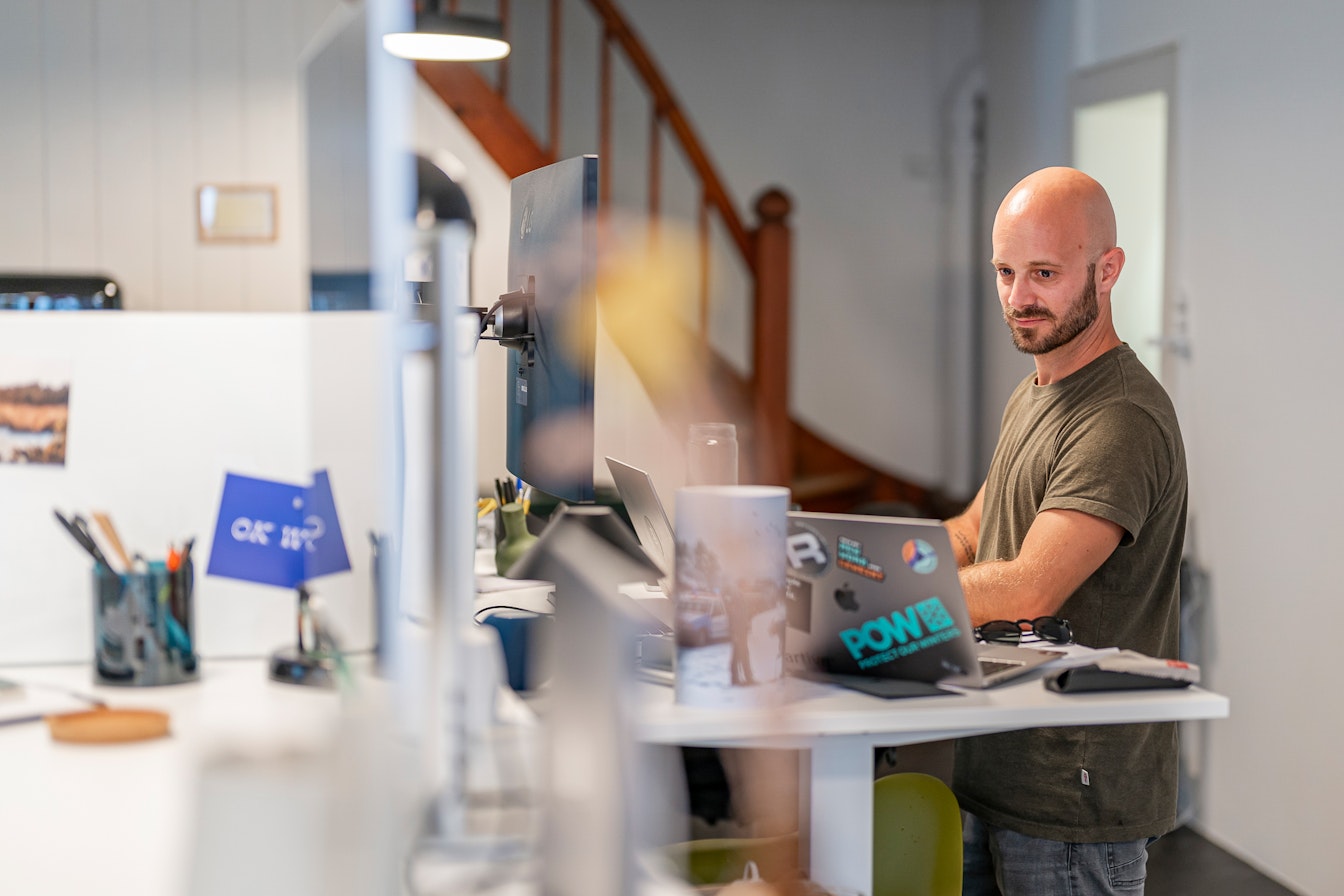 Bei smartive verwenden wir für Webseiten ausschliesslich Headless Content Management Systeme Ein Mann arbeitet an einem Stehpult in einem Büro und schaut auf einen Computermonitor. Er hat einen Laptop mit verschiedenen Aufklebern darauf und im Hintergrund sind Büromaterialien zu sehen.