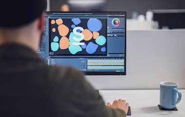 Eine Person arbeitet mithilfe einer Software auf einem Desktop-Computer an einem Grafikdesignprojekt. Auf dem Schreibtisch neben dem Monitor steht eine blaue Tasse.