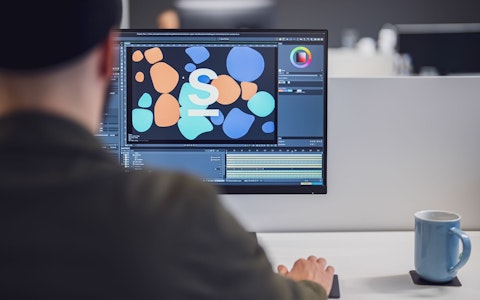Eine Person arbeitet mithilfe einer Software auf einem Desktop-Computer an einem Grafikdesignprojekt. Auf dem Schreibtisch neben dem Monitor steht eine blaue Tasse.