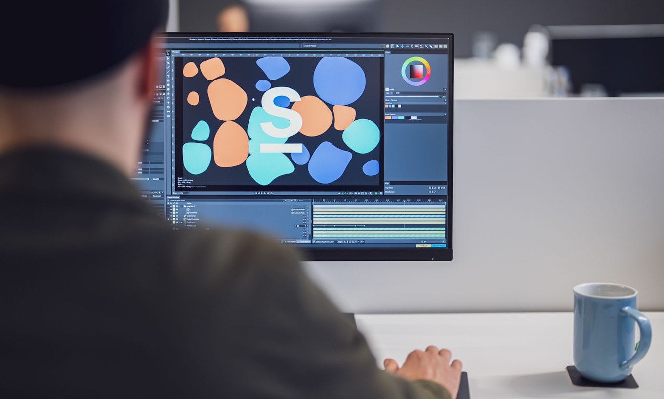 Eine Person arbeitet mithilfe einer Software auf einem Desktop-Computer an einem Grafikdesignprojekt. Auf dem Schreibtisch neben dem Monitor steht eine blaue Tasse.