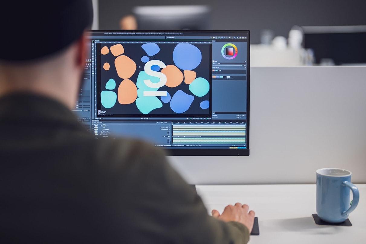 Eine Person arbeitet mithilfe einer Software auf einem Desktop-Computer an einem Grafikdesignprojekt. Auf dem Schreibtisch neben dem Monitor steht eine blaue Tasse.