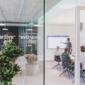 Ein Mann steht und präsentiert drei sitzenden Kollegen in einem modernen Büro mit Glaswänden und Grünpflanzen Informationen auf einem Bildschirm. An der Wand hinter ihnen ist das Wort „smartive“ zu sehen.