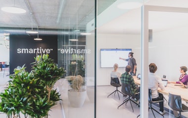 Ein Mann steht und präsentiert drei sitzenden Kollegen in einem modernen Büro mit Glaswänden und Grünpflanzen Informationen auf einem Bildschirm. An der Wand hinter ihnen ist das Wort „smartive“ zu sehen.