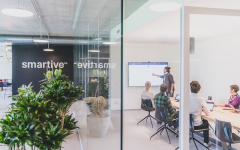 Ein Mann steht und präsentiert drei sitzenden Kollegen in einem modernen Büro mit Glaswänden und Grünpflanzen Informationen auf einem Bildschirm. An der Wand hinter ihnen ist das Wort „smartive“ zu sehen.