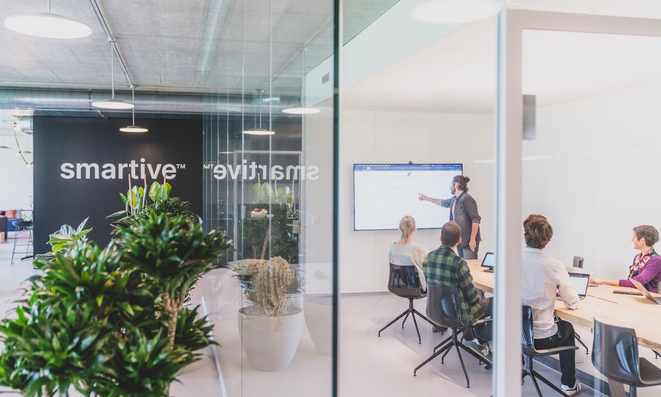 Ein Mann steht und präsentiert drei sitzenden Kollegen in einem modernen Büro mit Glaswänden und Grünpflanzen Informationen auf einem Bildschirm. An der Wand hinter ihnen ist das Wort „smartive“ zu sehen.